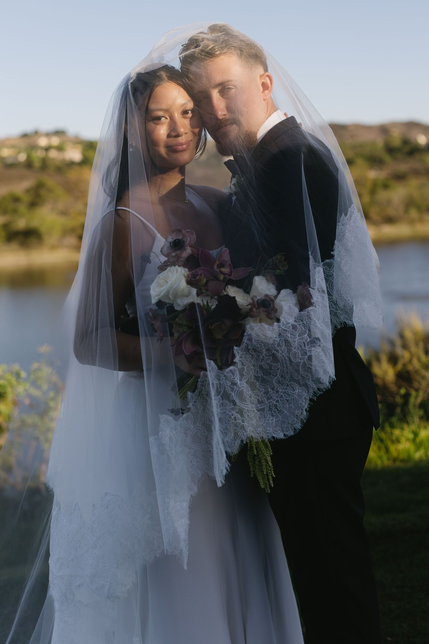 A romantic wedding portrait captured at sunset by a lake with a lace veil flowing in the breeze.