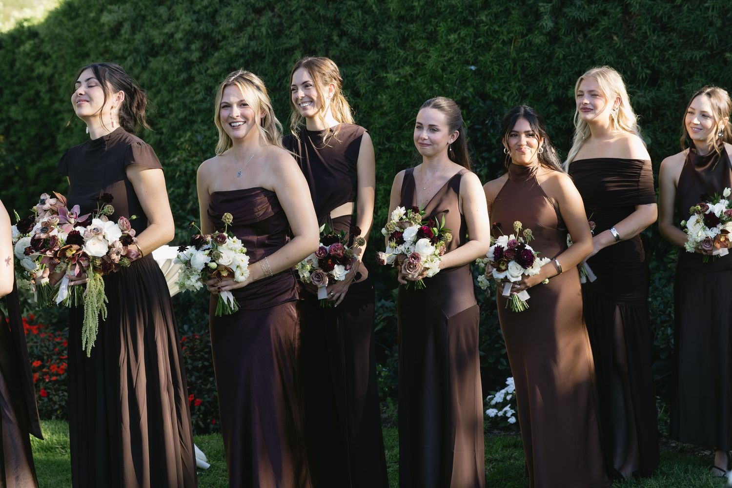 Bridesmaids in matching brown dresses stand in line holding bouquets at outdoor wedding.