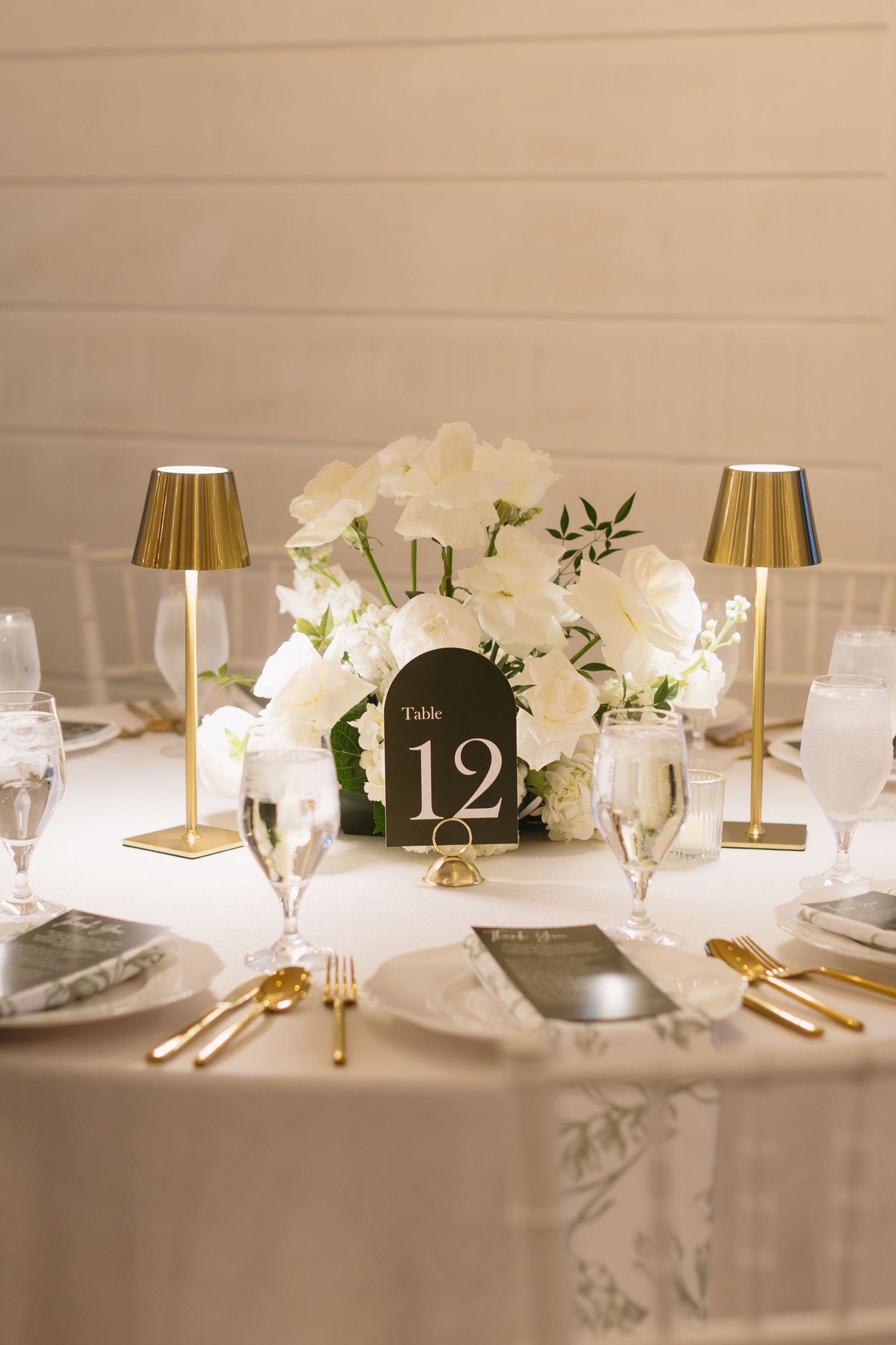 Elegant wedding reception table setting with white flowers, gold lamps, and table numbers in classic decor.