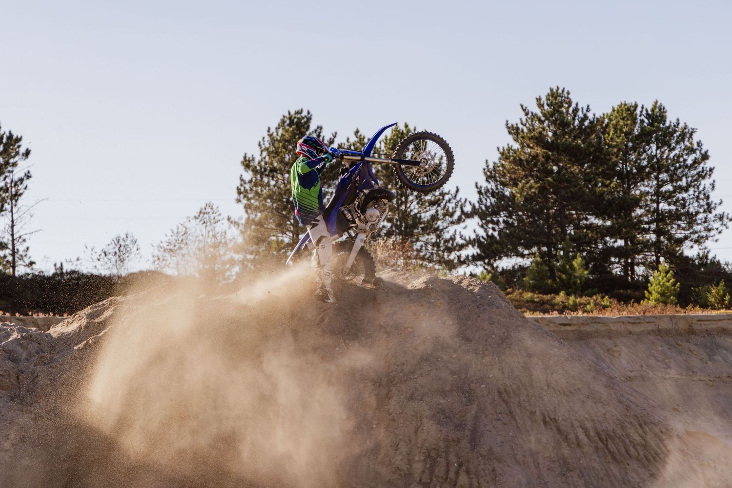 Creative portrait dirtbike photoshoot. Rider is holding the bike in a wheelie on top of a dirt pile. Sand is blowing in the wind.