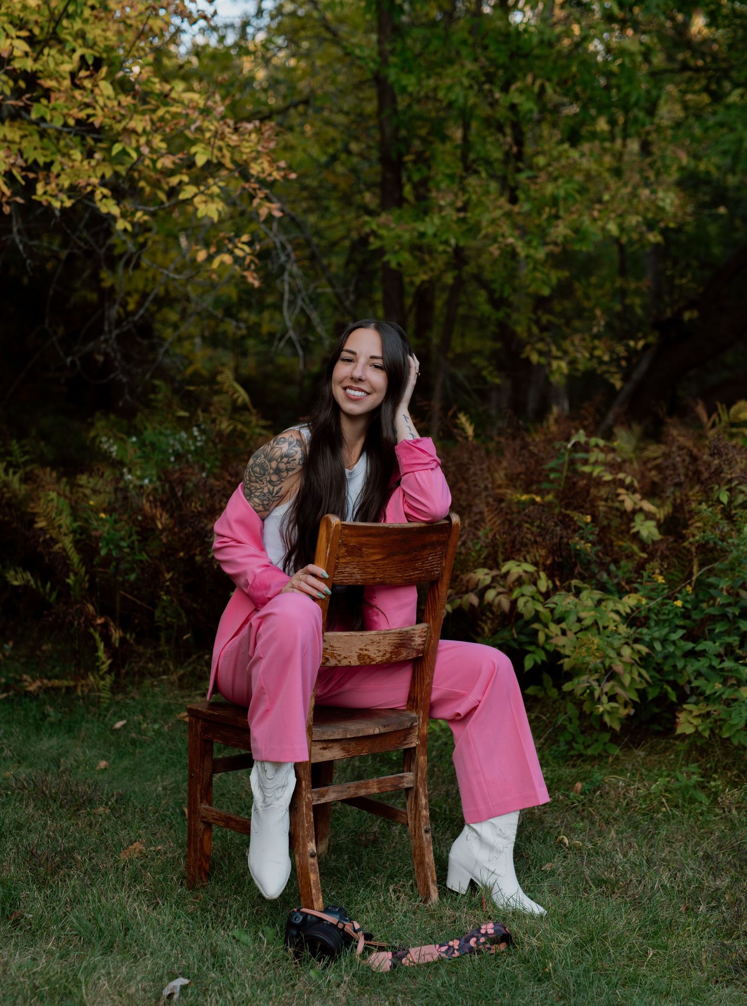 Sidney smiling at the camera, wearing a hot pink suit with white cowboy boots, sitting backwards in a rustic chair in the forest.