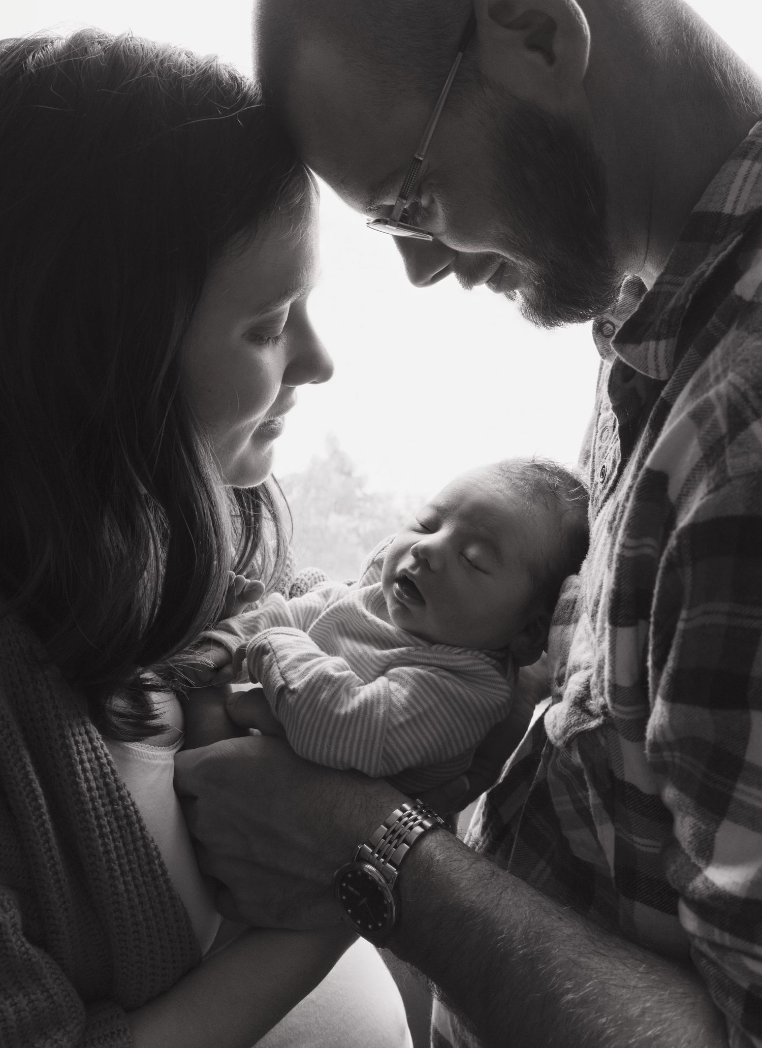 Lifestyle newborn photoshoot. Parents holding baby between them, looking at her.