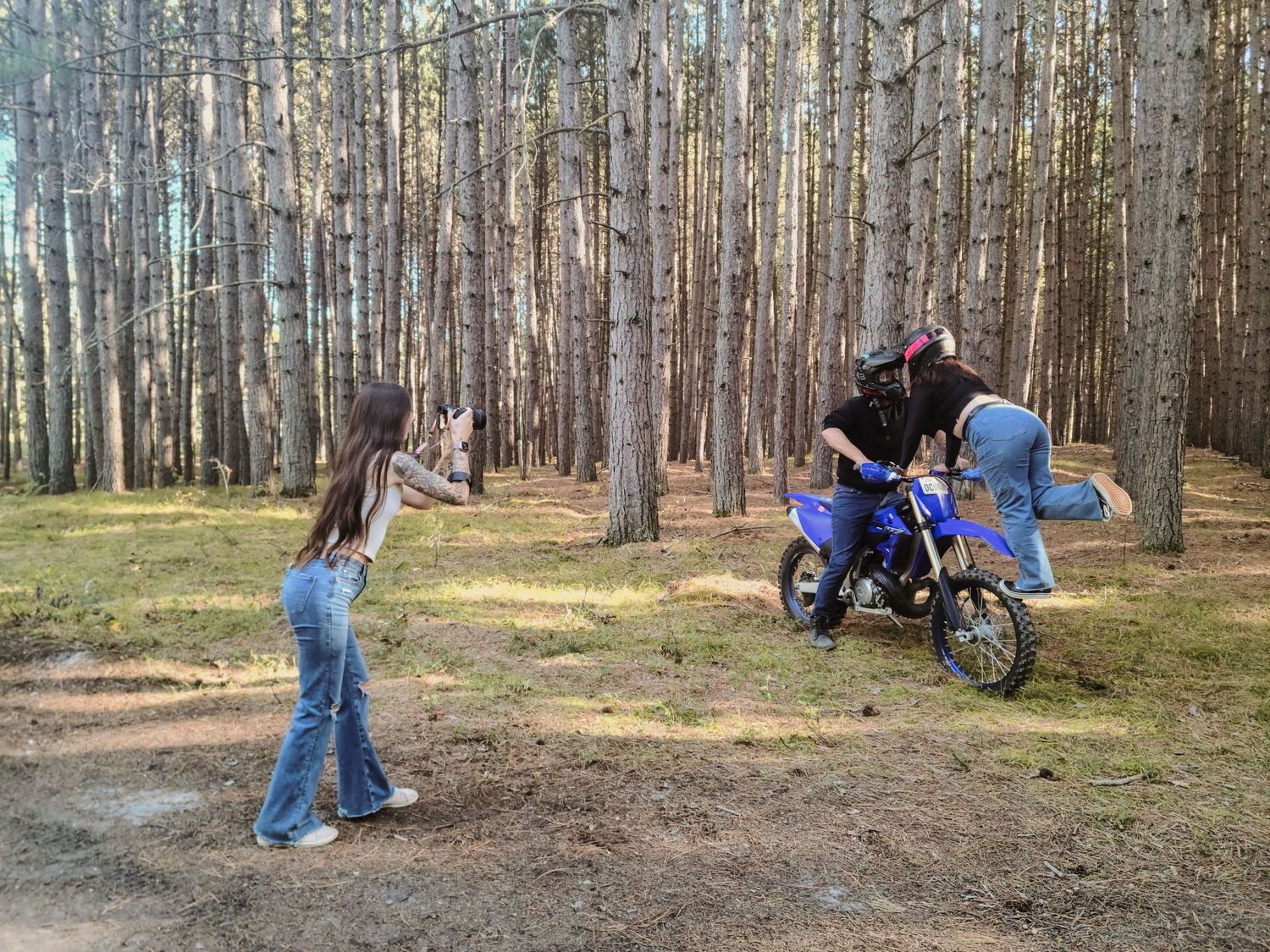 Sidney in the forest, photographing a couple. The man is sitting on a dirtbike, and the woman standing on the front, leaning in toward him.