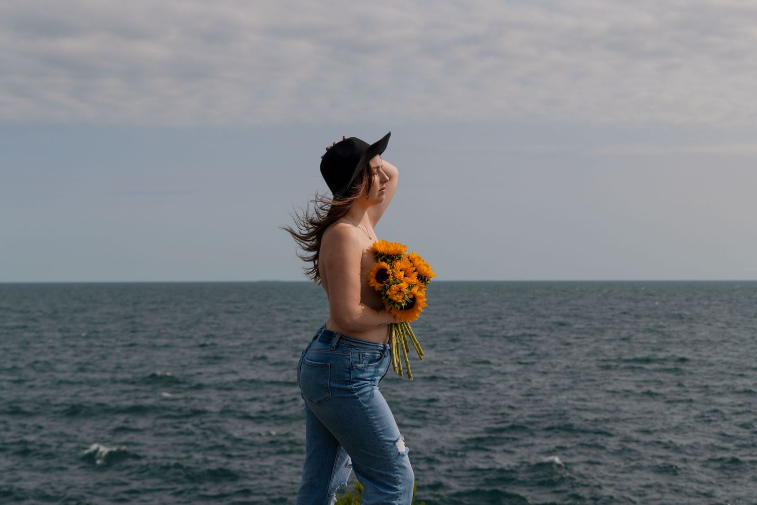A woman standing in front of Georgian Bay, her hair blowing in the wind. She is wearing jeans and holding sunflowers in front of her chest.