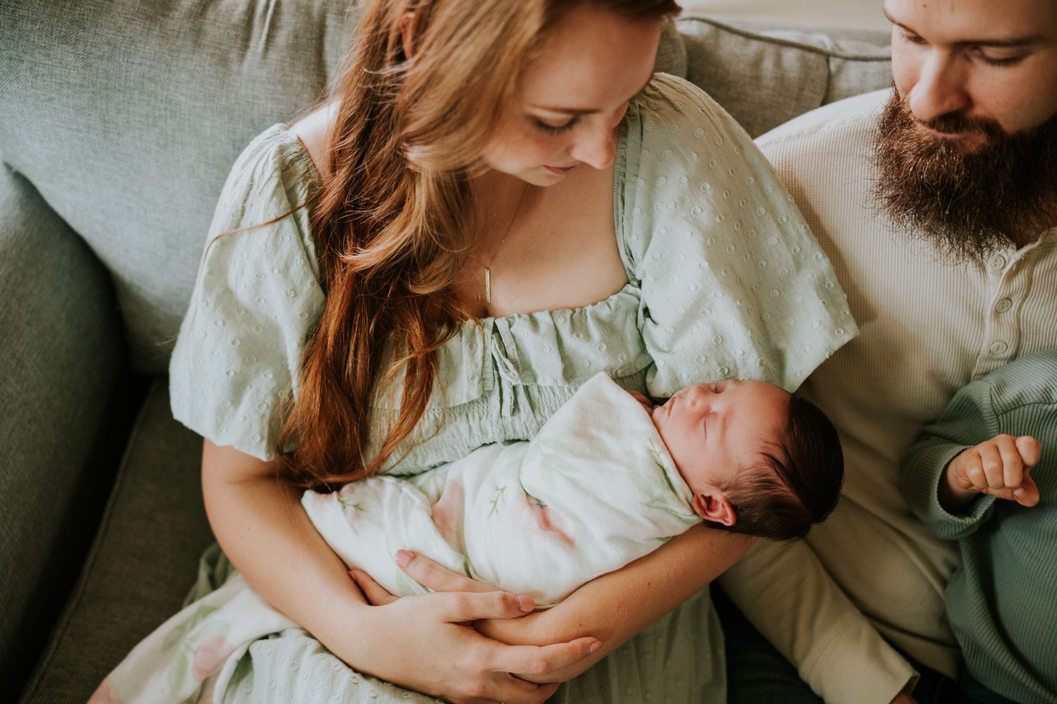 Smiling mother in sage green dress gazes at her swaddled newborn baby while father looks on.