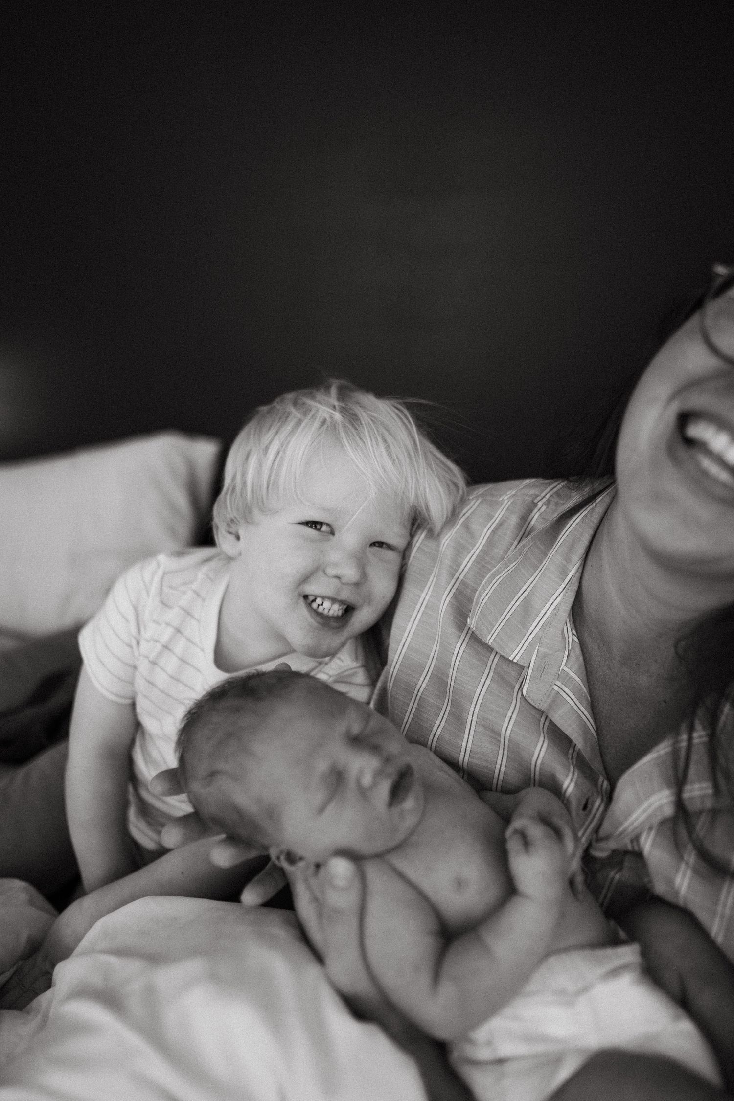 Black and white photo of laughing mother with toddler and newborn baby on bed.