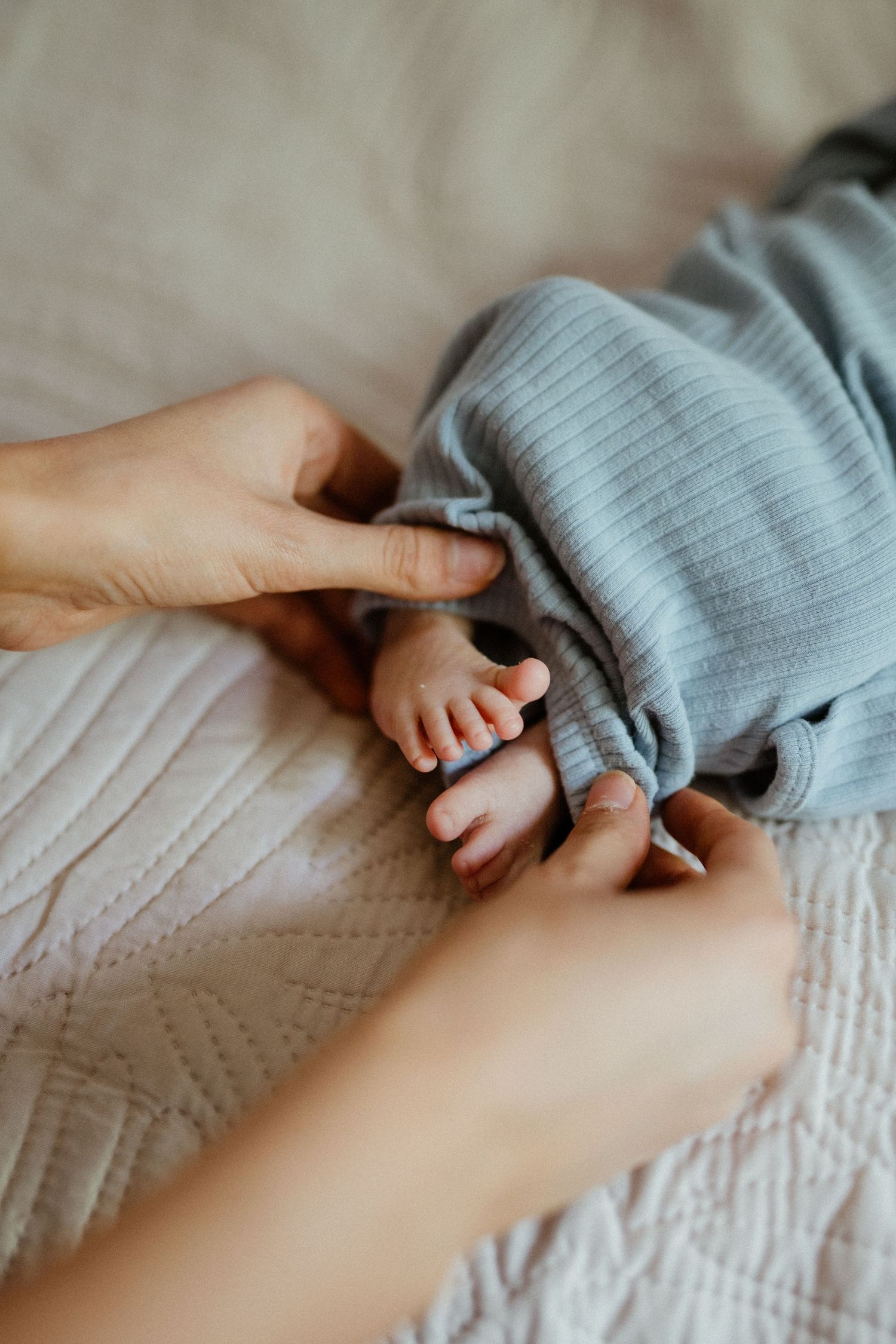 Parent gently holding tiny newborn baby feet dressed in blue ribbed pants on white bedding.