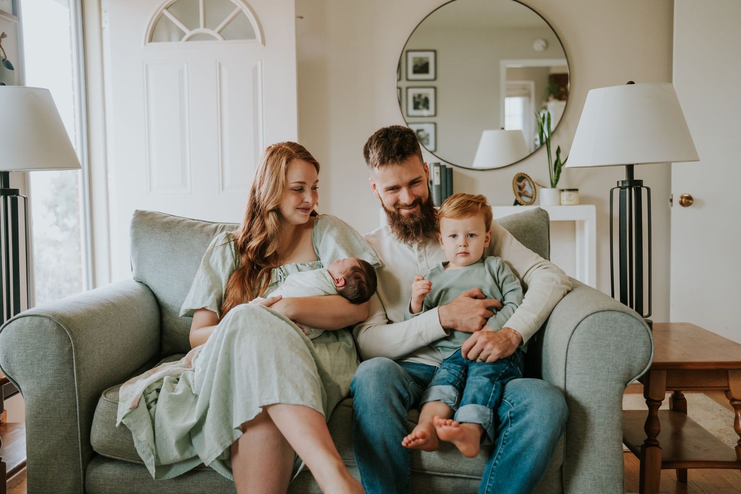 Happy young family with bearded father, mother in green dress, toddler, and newborn on a gray couch.
