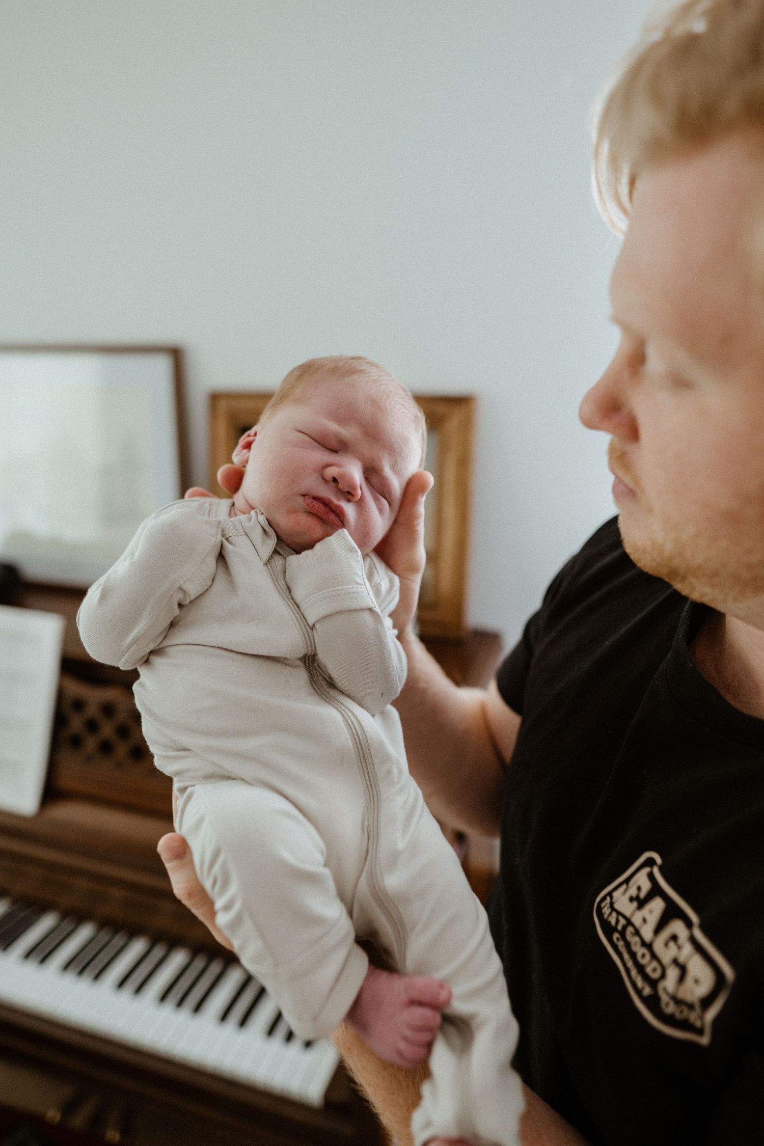 Blonde father in black t-shirt gently holds sleeping newborn baby near an upright piano.
