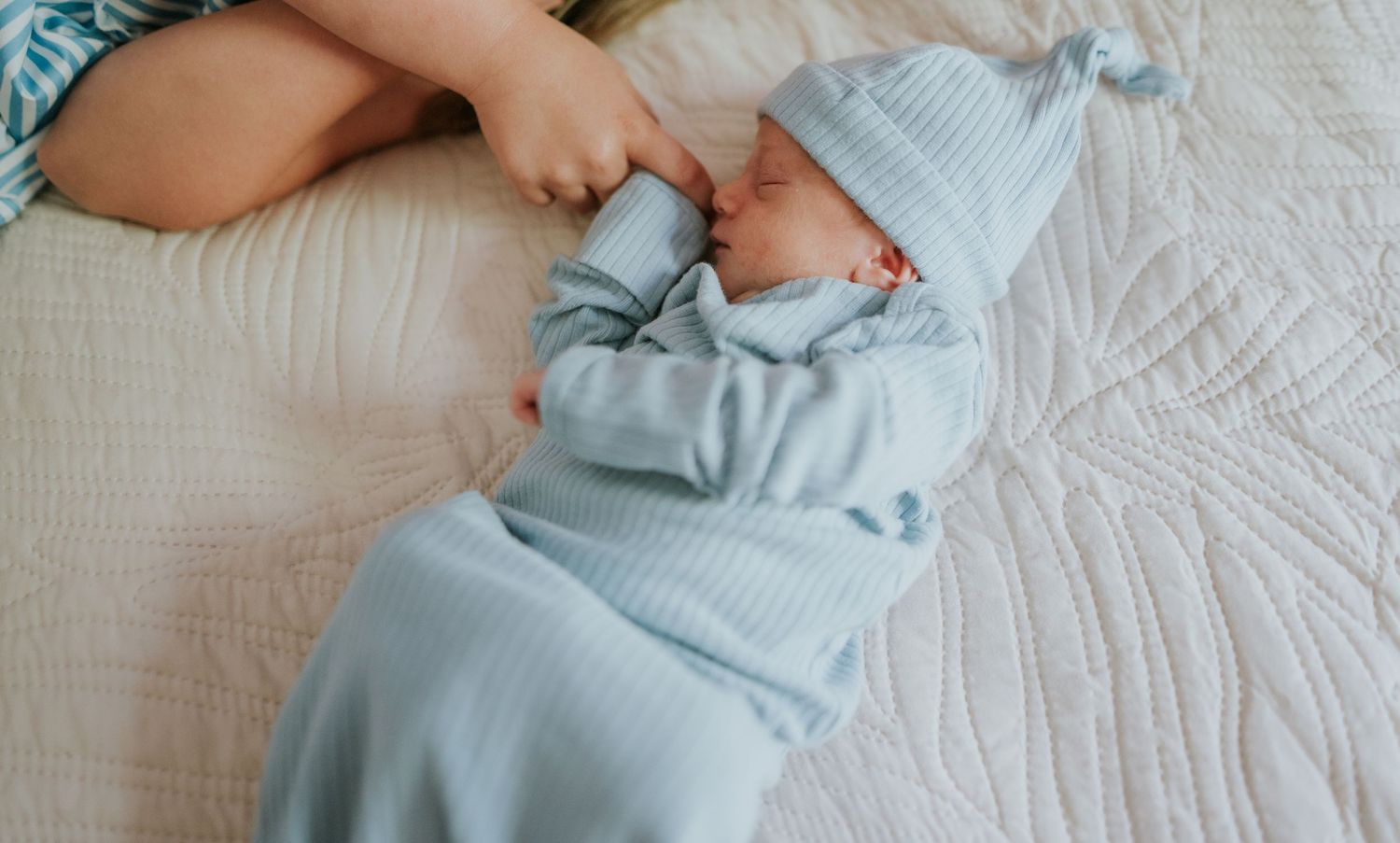 Sleeping newborn baby in light blue knit outfit and hat lying on white quilted bedding beside sibling.
