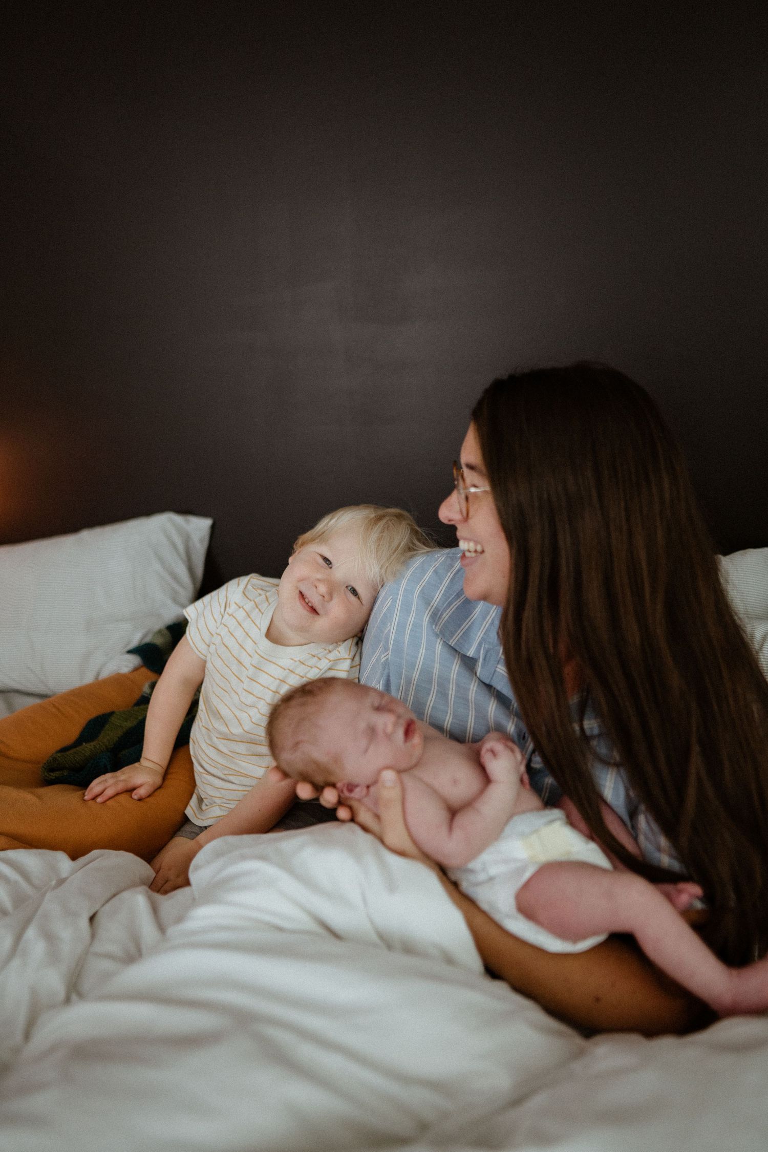 Brunette mother in blue striped shirt smiles at laughing toddler with newborn baby on white bed.
