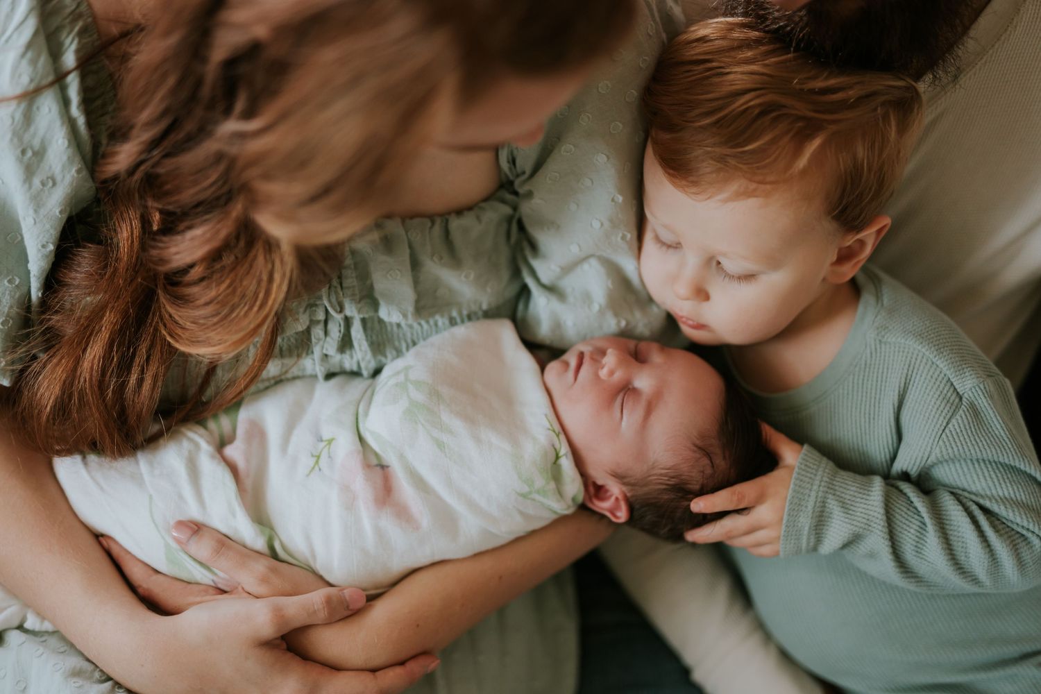 Mother holds swaddled newborn while red-haired toddler leans in to gently kiss the baby.