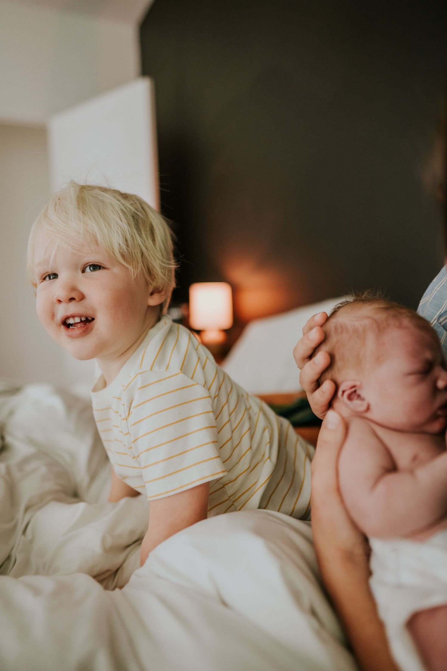 Smiling blonde toddler in striped shirt sits on bed next to parent holding a newborn baby.