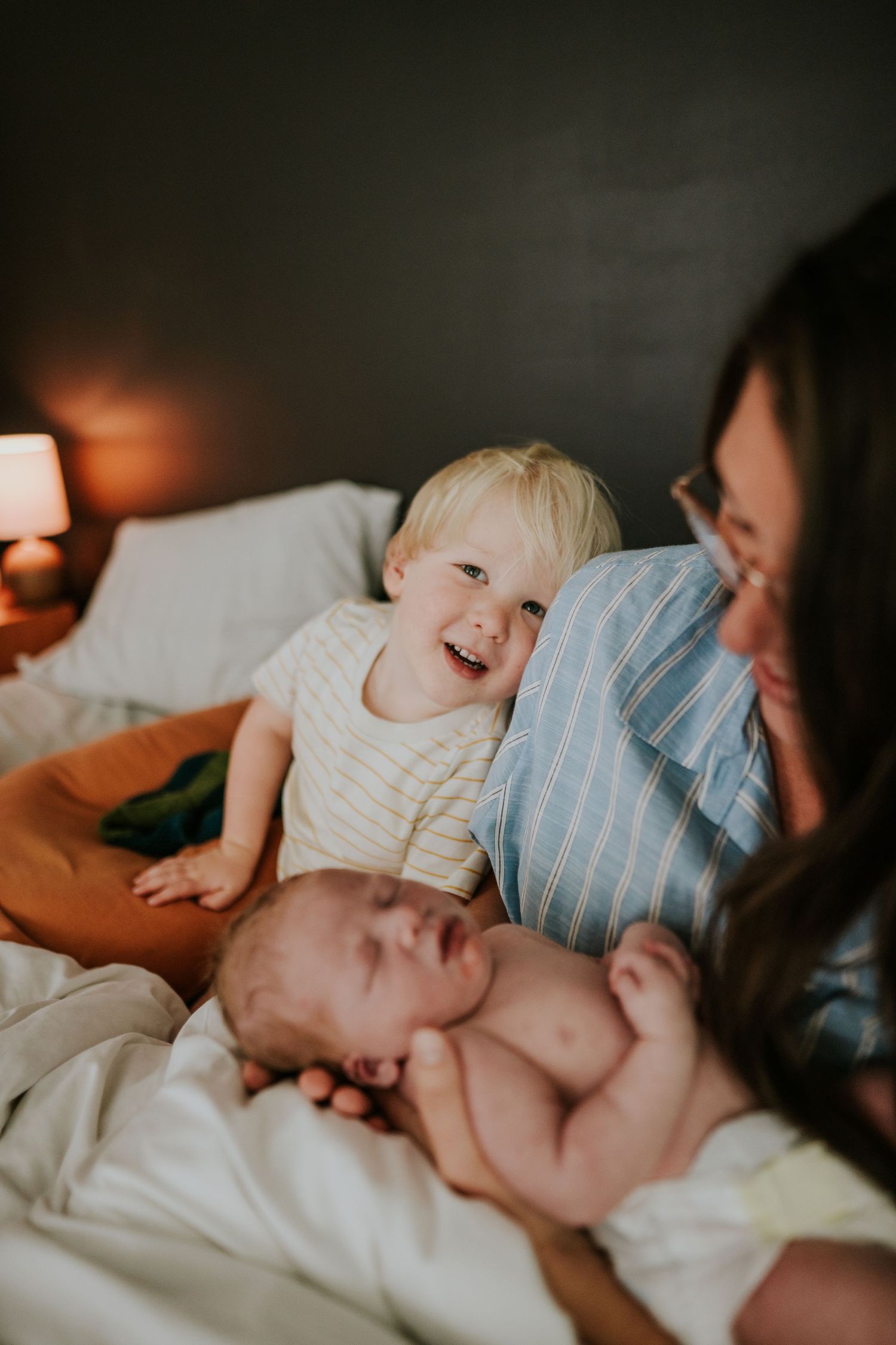 Cheerful toddler leans on smiling mother holding newborn baby on white bed with warm lamp glow.