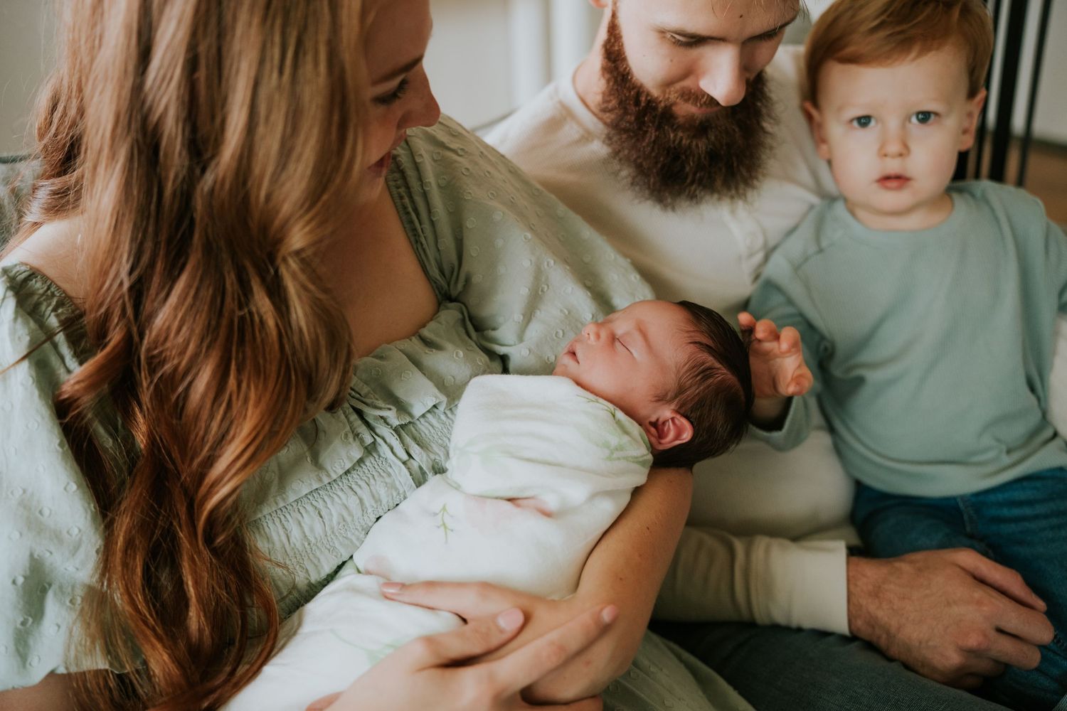 Parents and red-haired toddler gaze lovingly at swaddled newborn baby on living room chair.