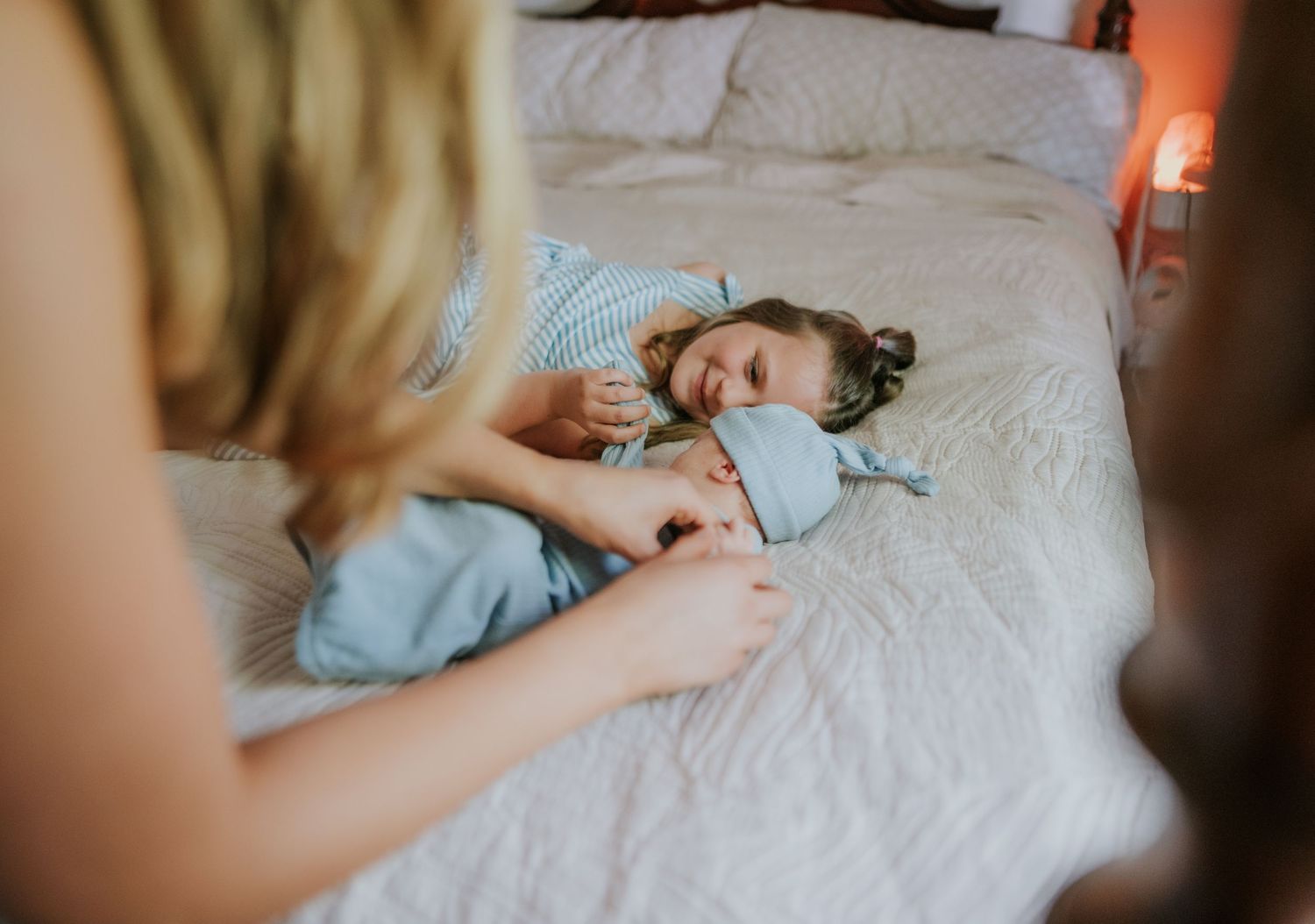 Young girl lying on white bed smiling while someone buttons her light blue pajamas in warm bedroom light.