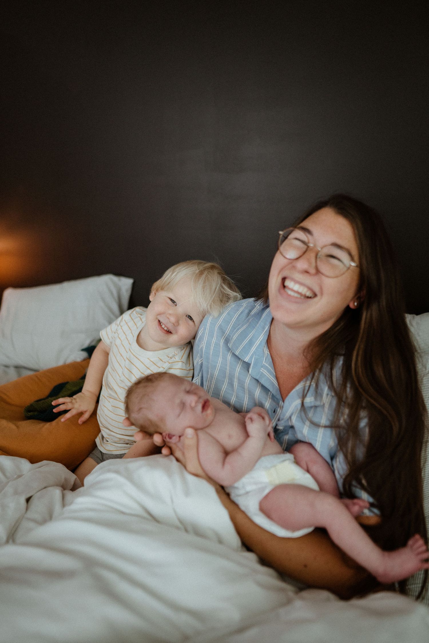 Smiling brunette mother with glasses laughs holding newborn while toddler poses beside her on bed.