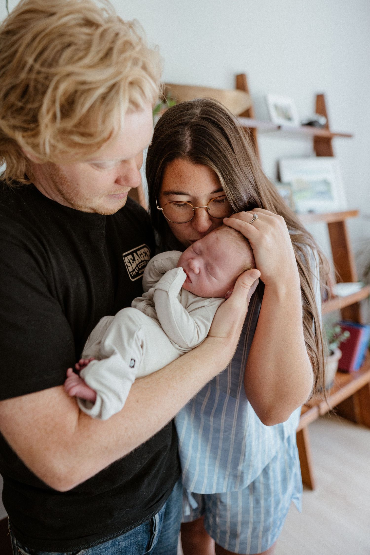 Parents with curly blonde and long dark hair tenderly kiss their sleeping newborn baby together.