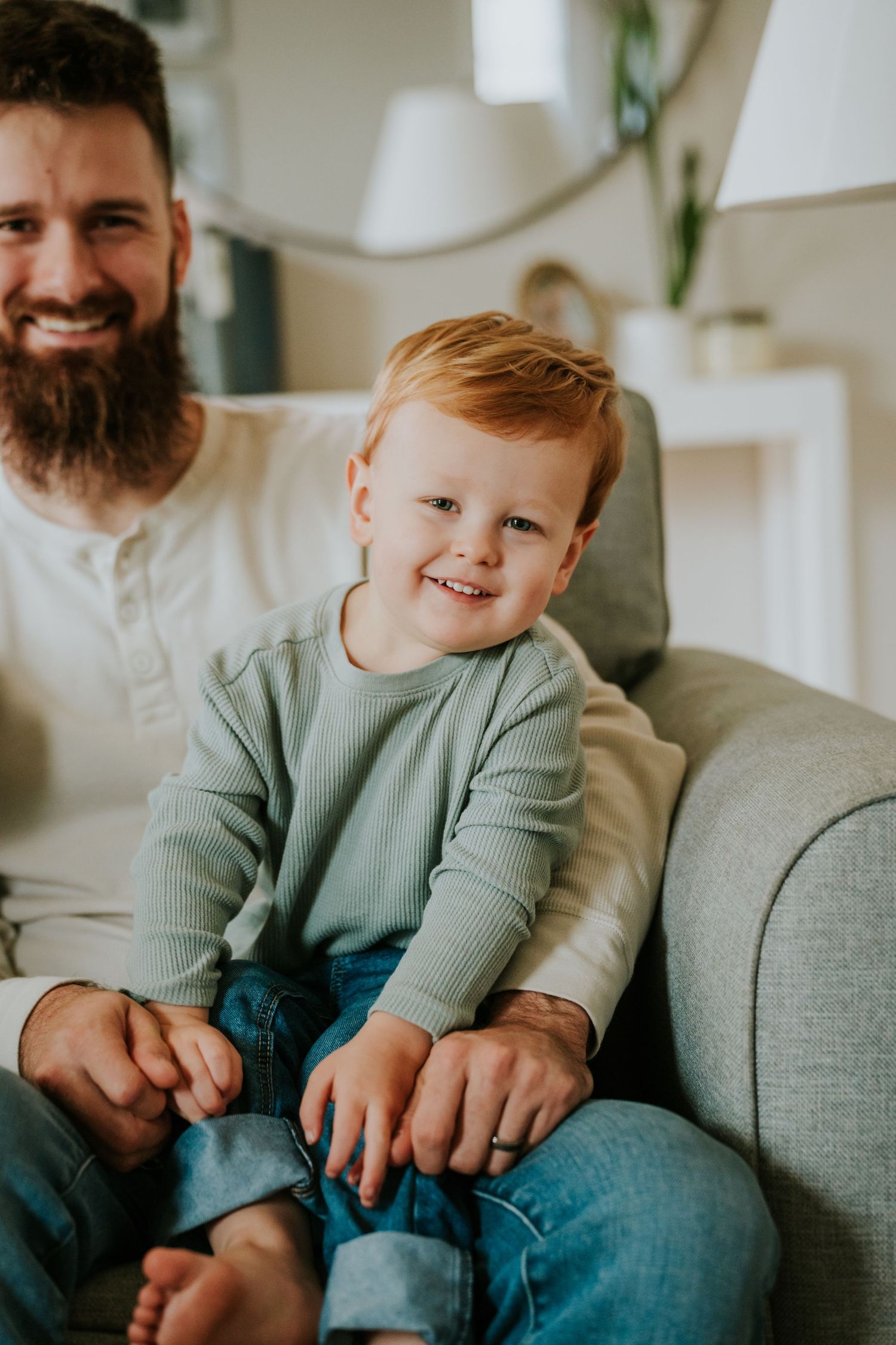 Smiling red-haired toddler boy sitting on a gray sofa with his bearded father in a bright home.