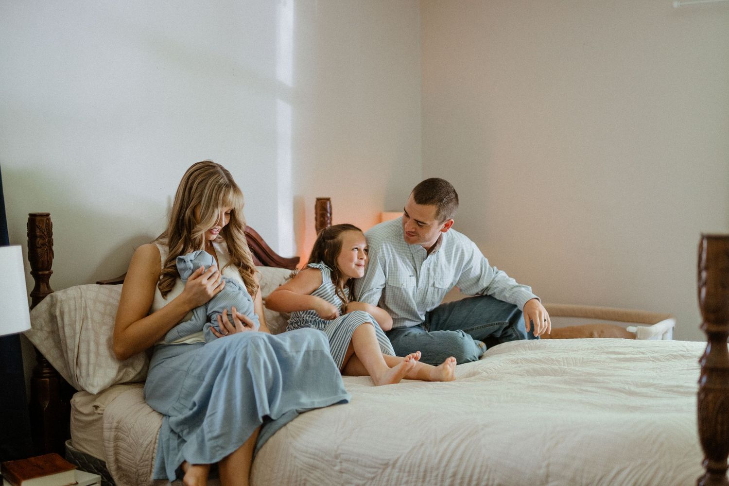 Parents and young daughter sit on bed admiring newborn baby in warmly lit bedroom setting.