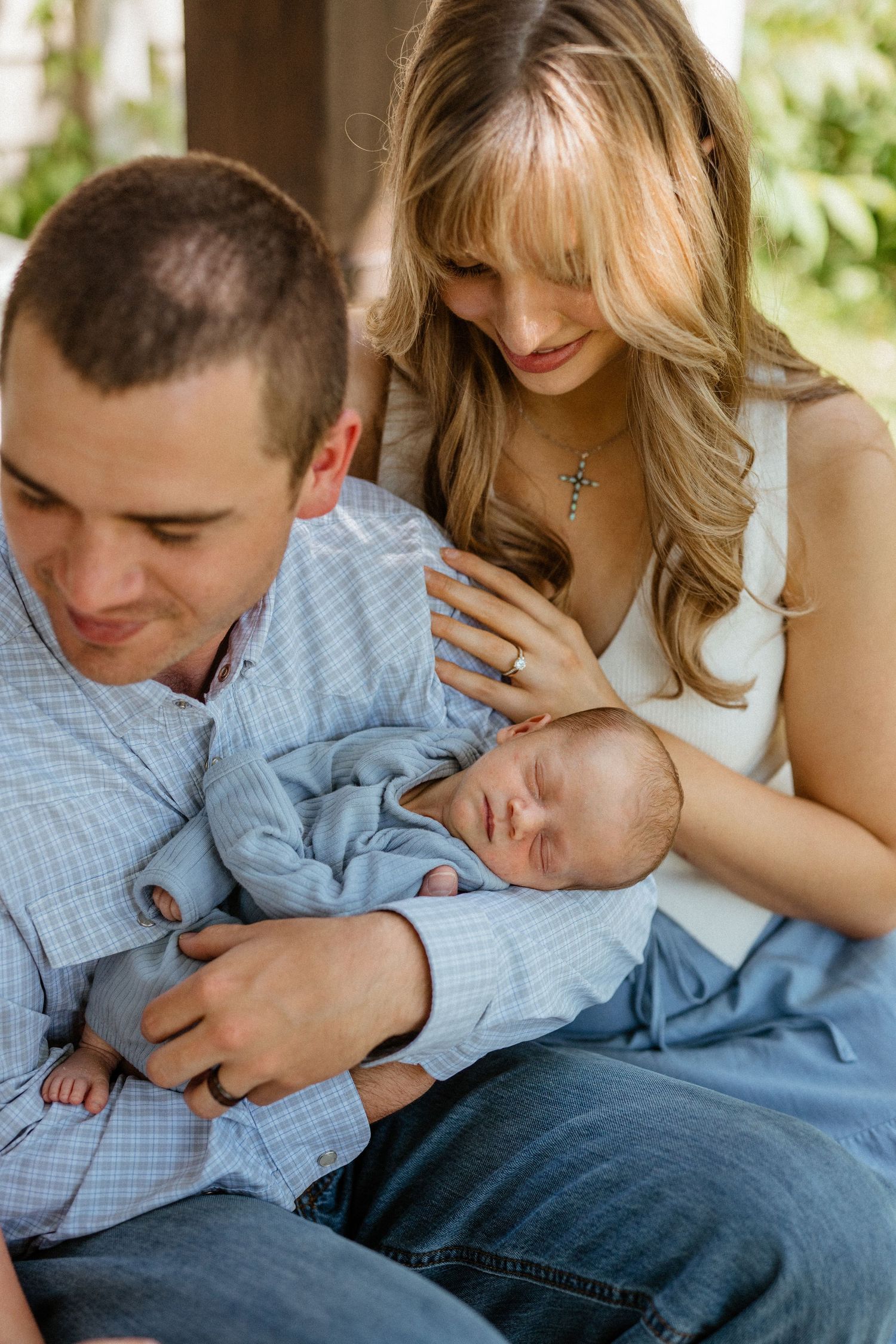 New parents in blue outfits tenderly hold sleeping newborn baby on porch.