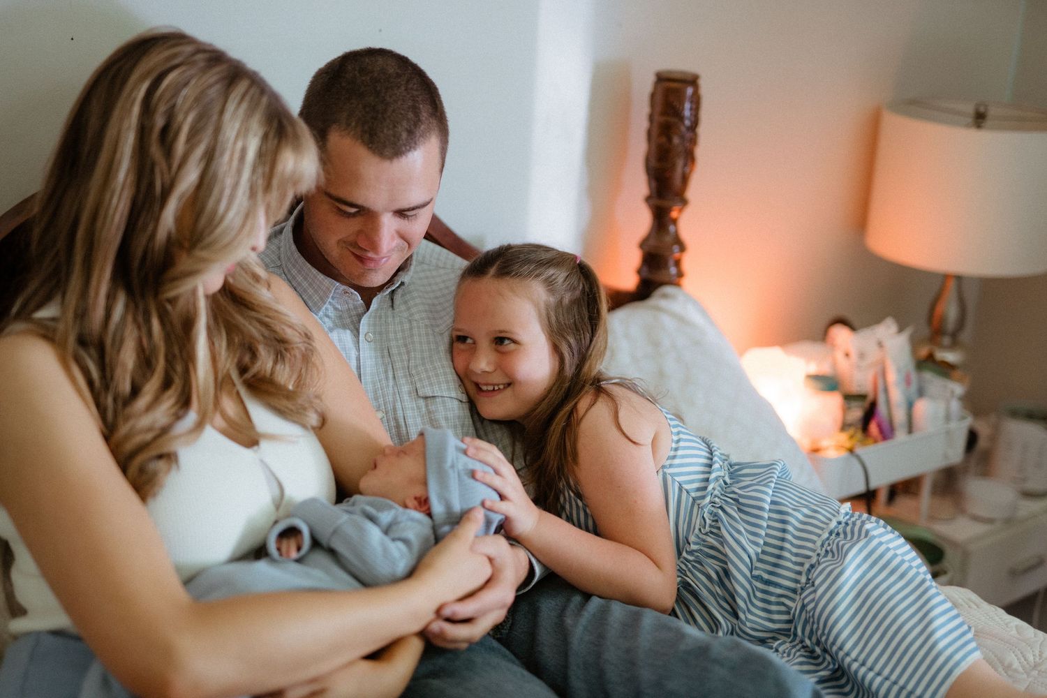 Young family of four smiling together on bed with newborn baby and excited older sister.