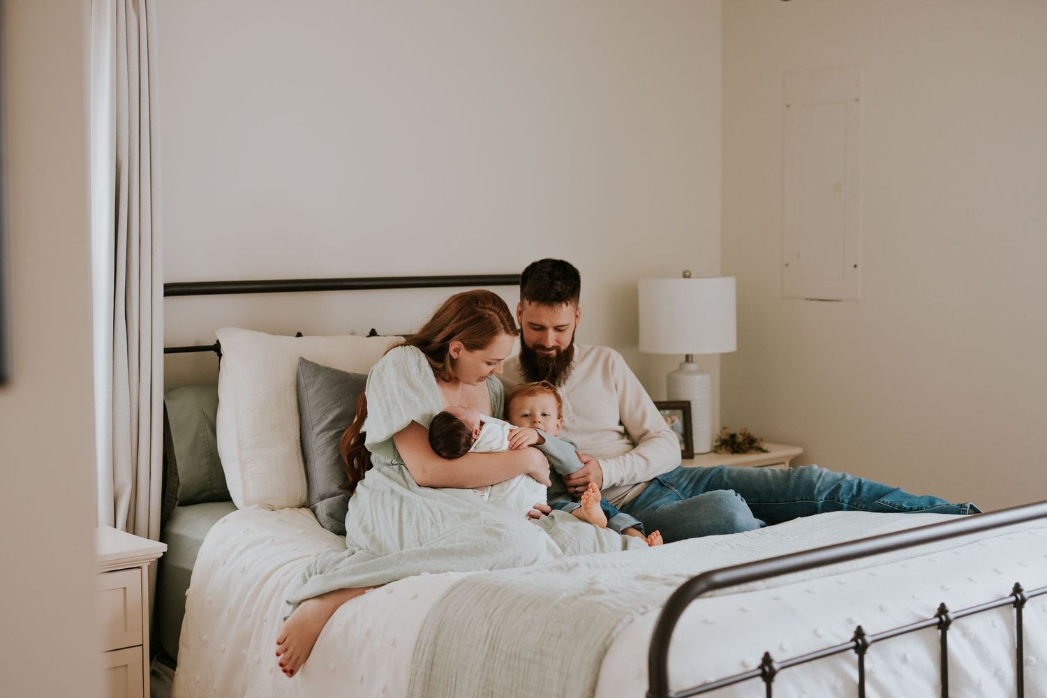Parents sitting on bed together lovingly attending to toddler and newborn baby.