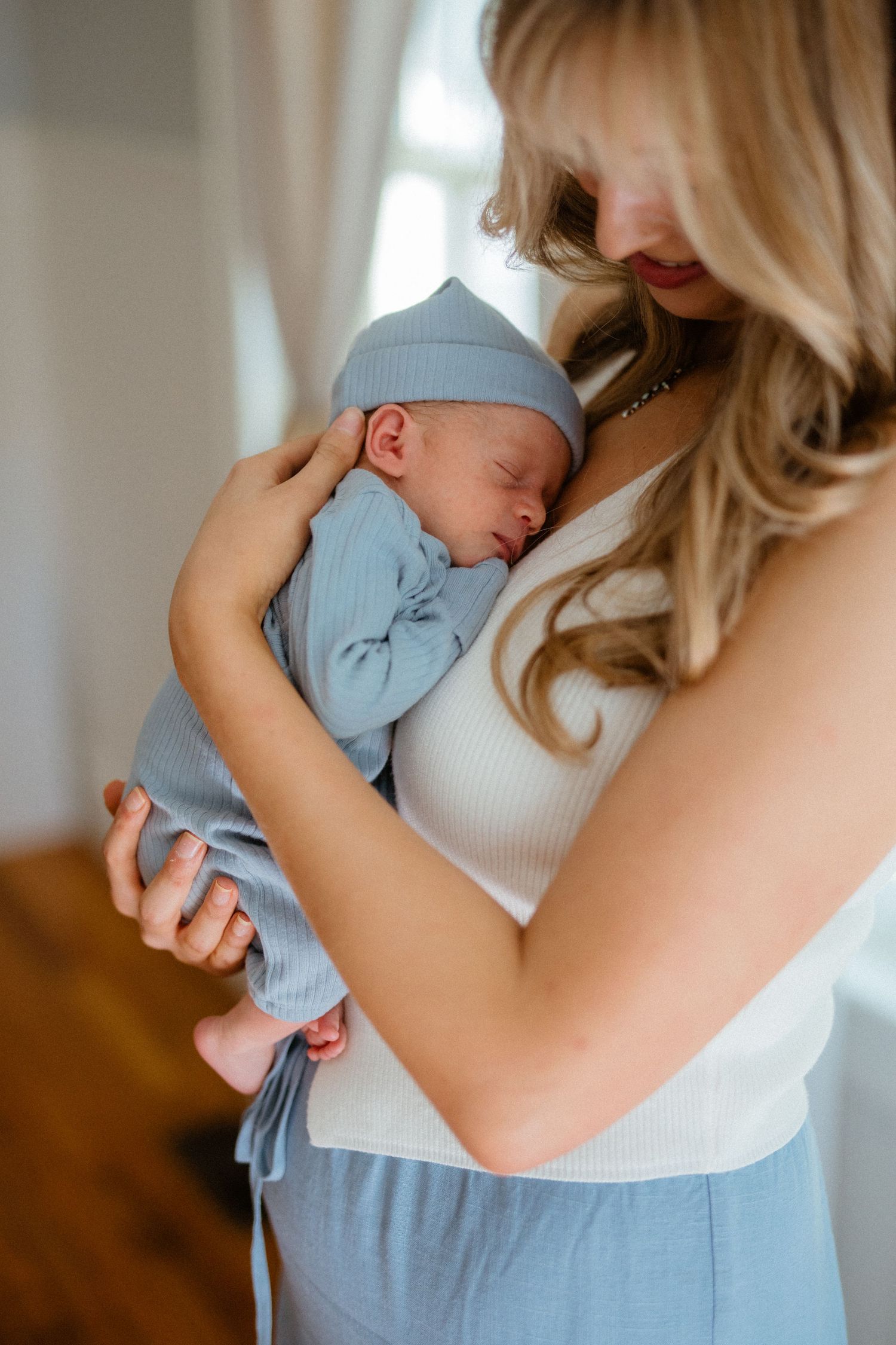 Blonde mother tenderly cradles her sleeping newborn wrapped in a soft blue swaddle and hat.