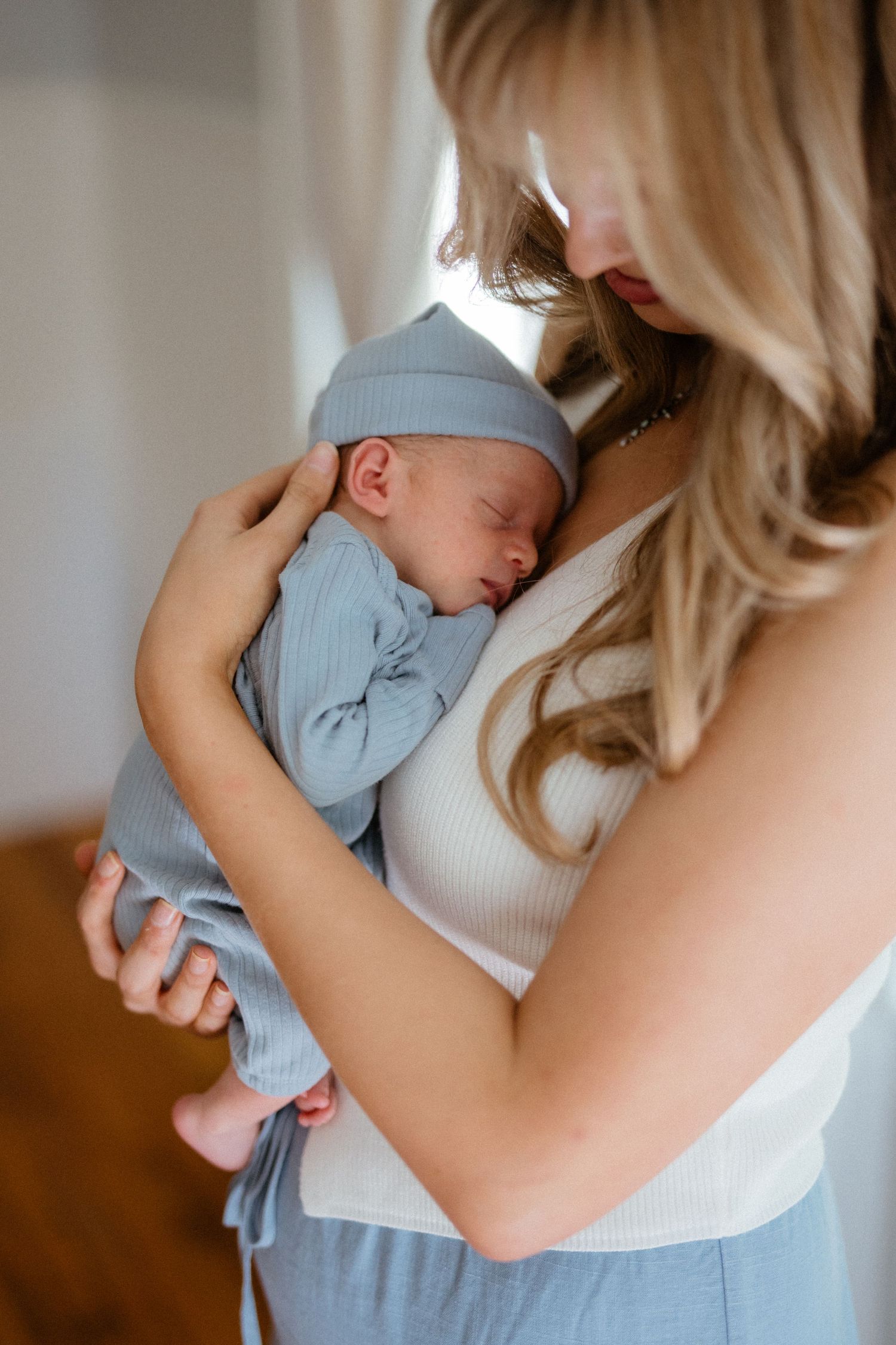 Blonde mother tenderly holds sleeping newborn dressed in a blue ribbed outfit and hat.