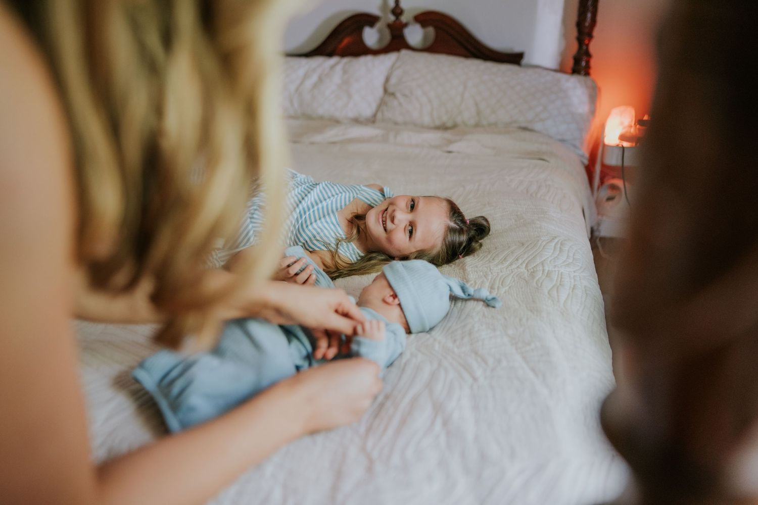 Laughing toddler lies on white bed while parents interact with them playfully.