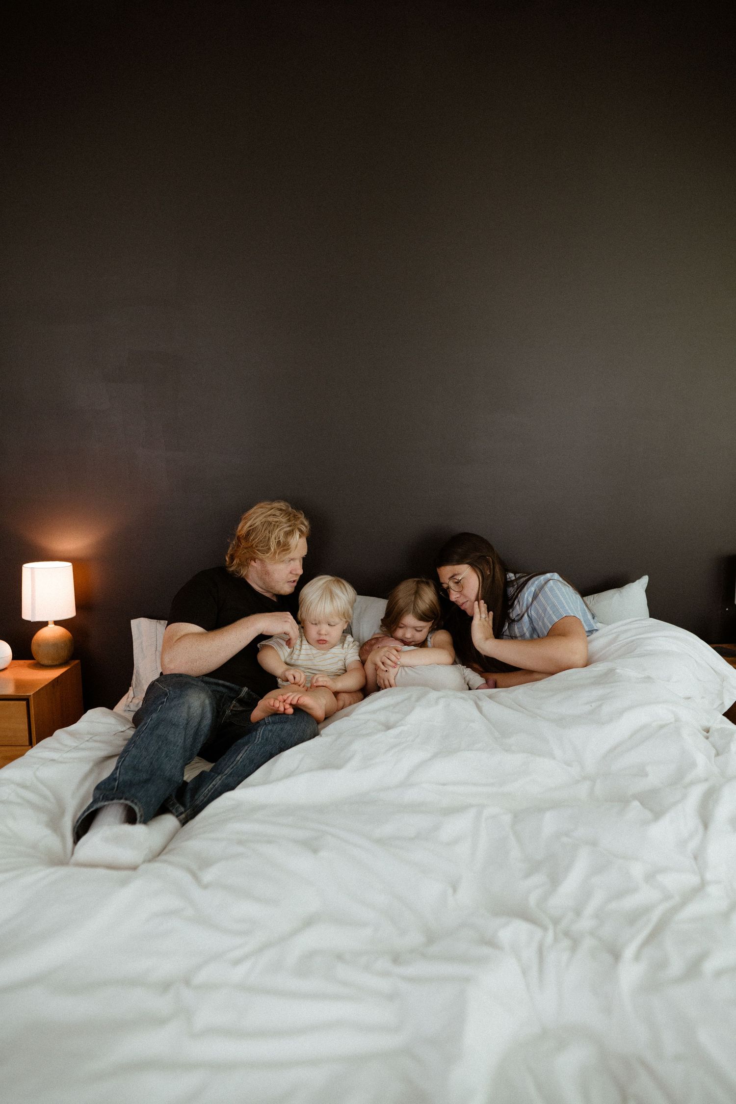 Family of four cuddles together on a white bed against a dark wall with a warm lamp glowing nearby.