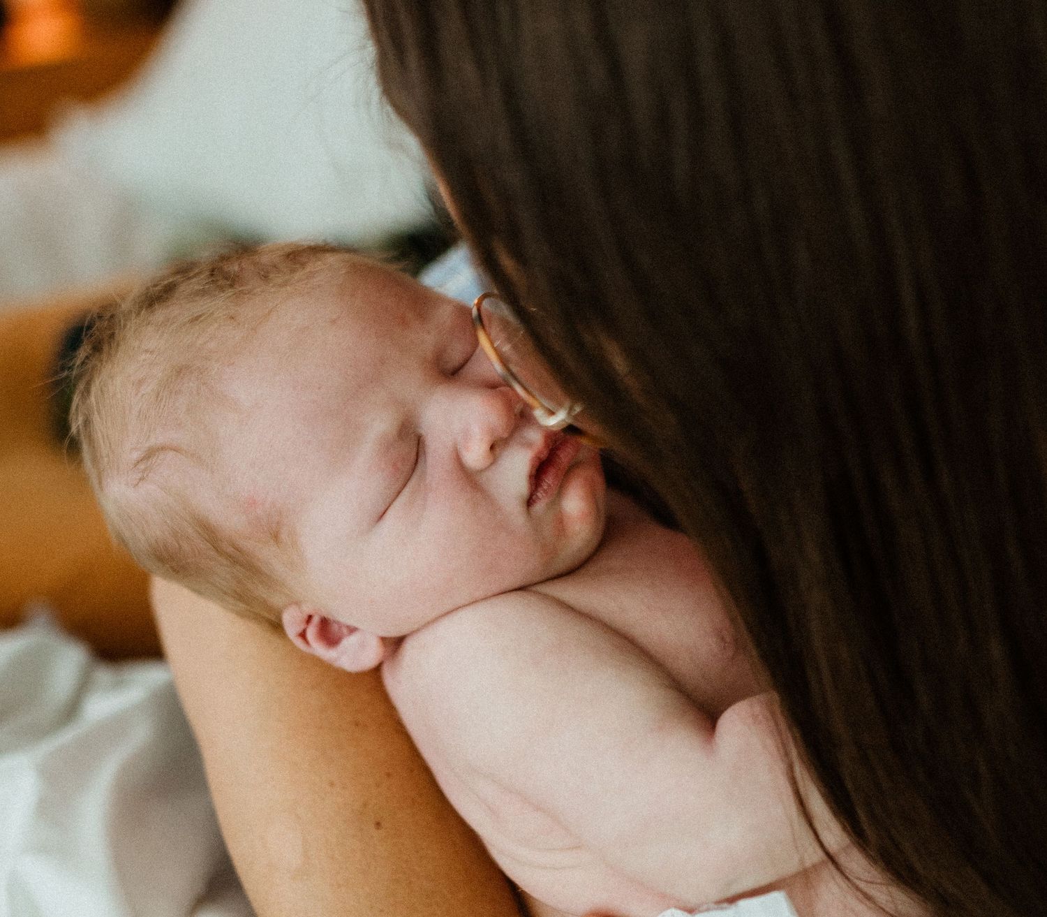 Mother with dark hair and glasses gently holds sleeping newborn baby skin-to-skin on her shoulder.