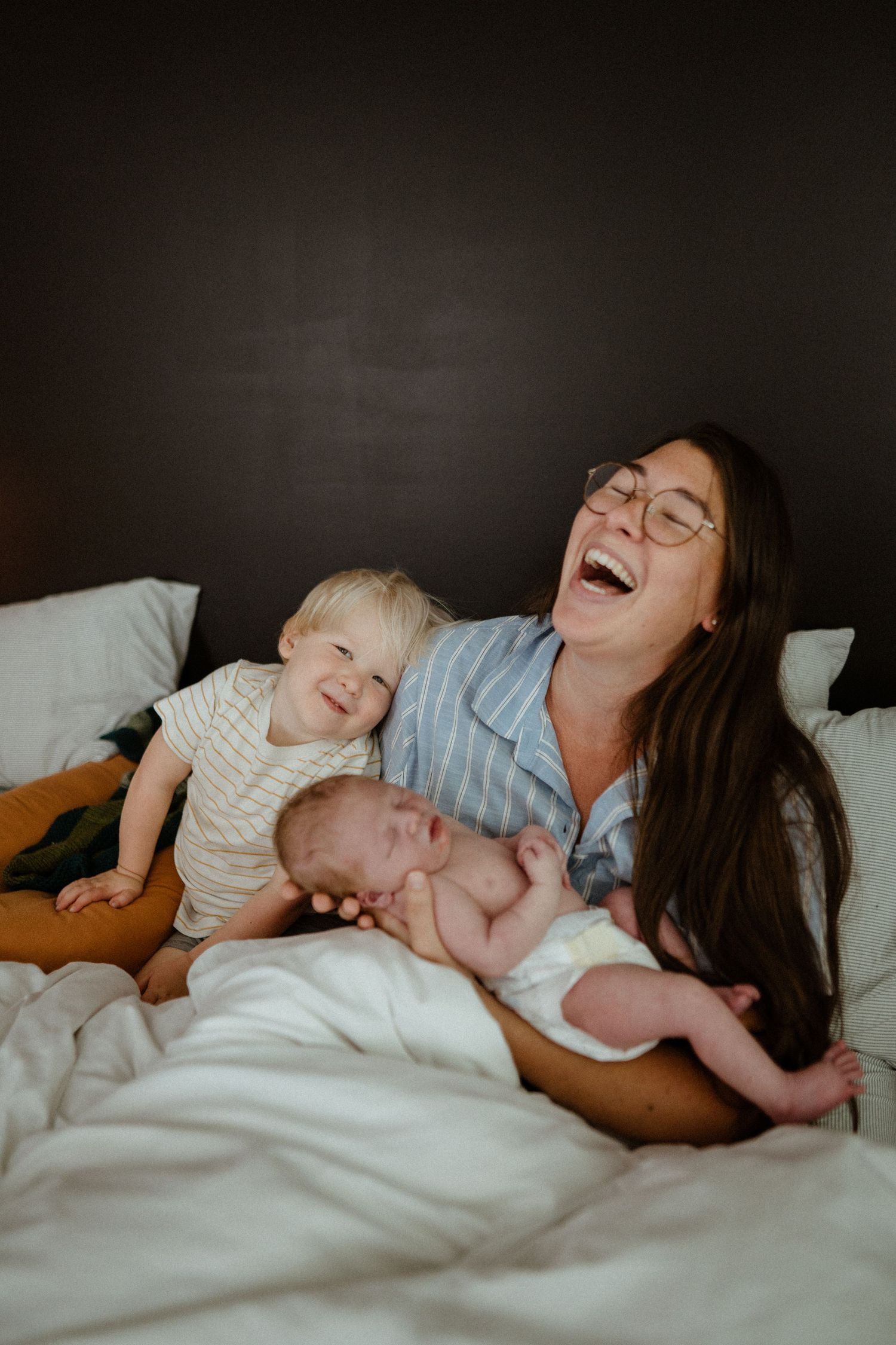 Laughing mother with glasses cuddles newborn and smiling toddler on white bed against dark wall.