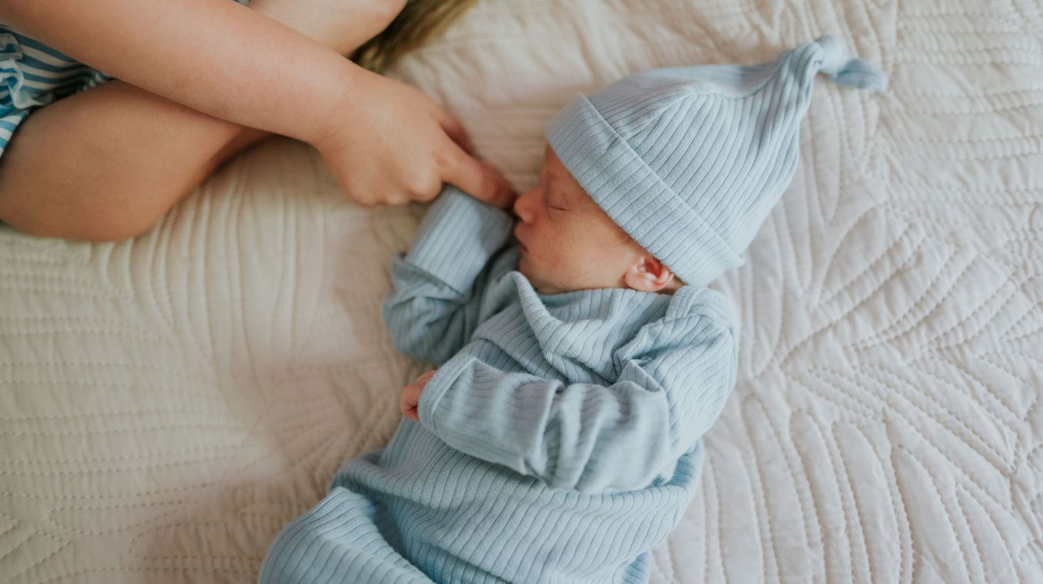 Overhead view of peaceful newborn in blue ribbed sleeper and knotted hat resting on white quilt.