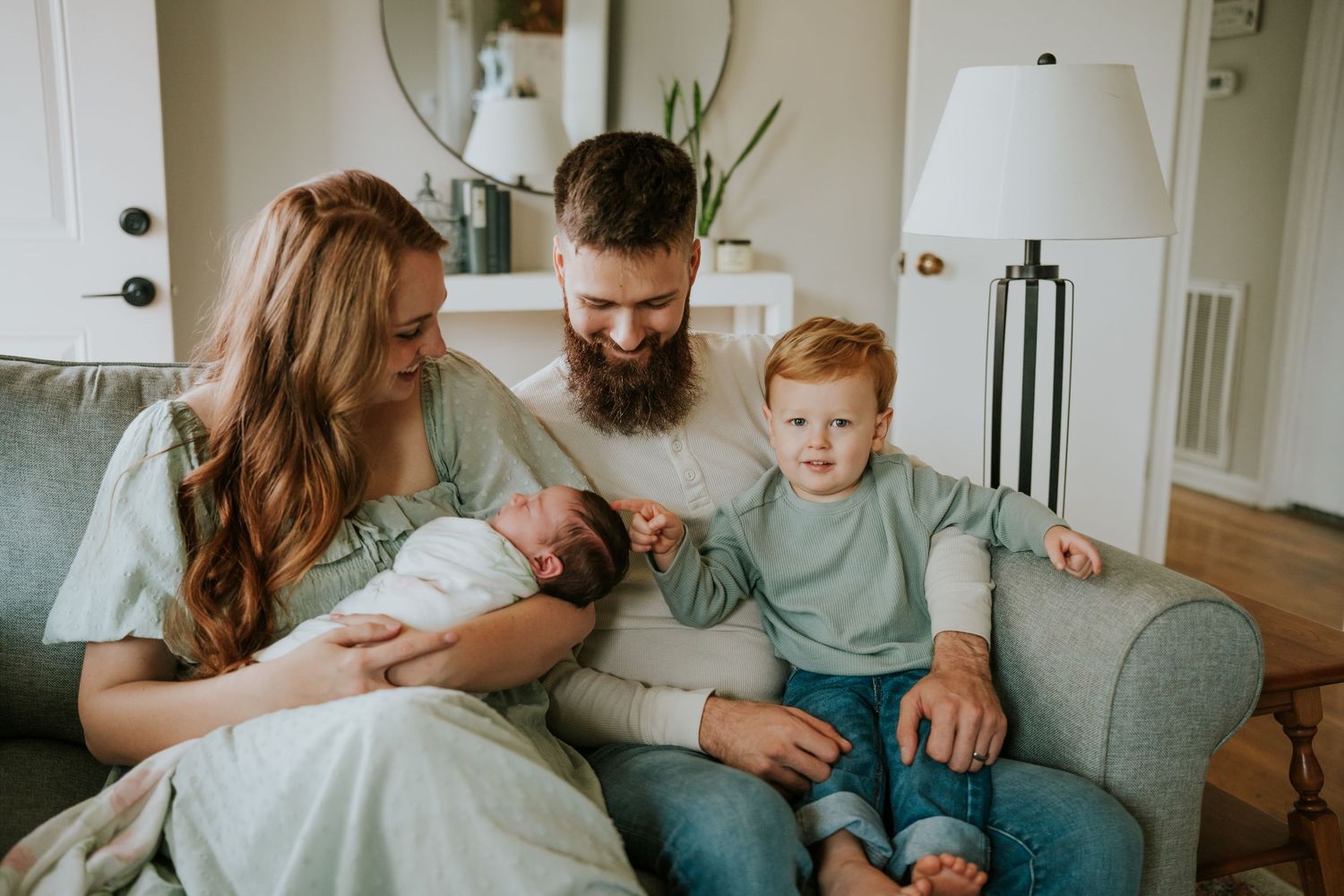 Smiling bearded father and mother with toddler son admire newborn baby on cozy armchair.