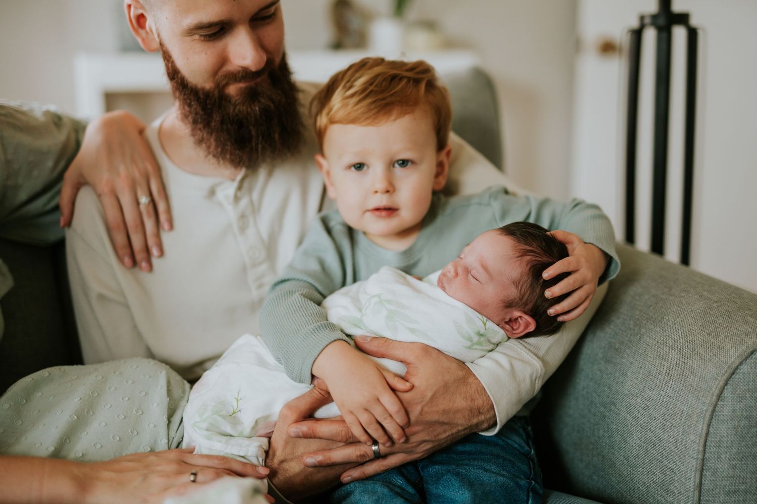 Bearded father holds sleeping newborn baby while red-haired toddler sits beside them on a sofa.