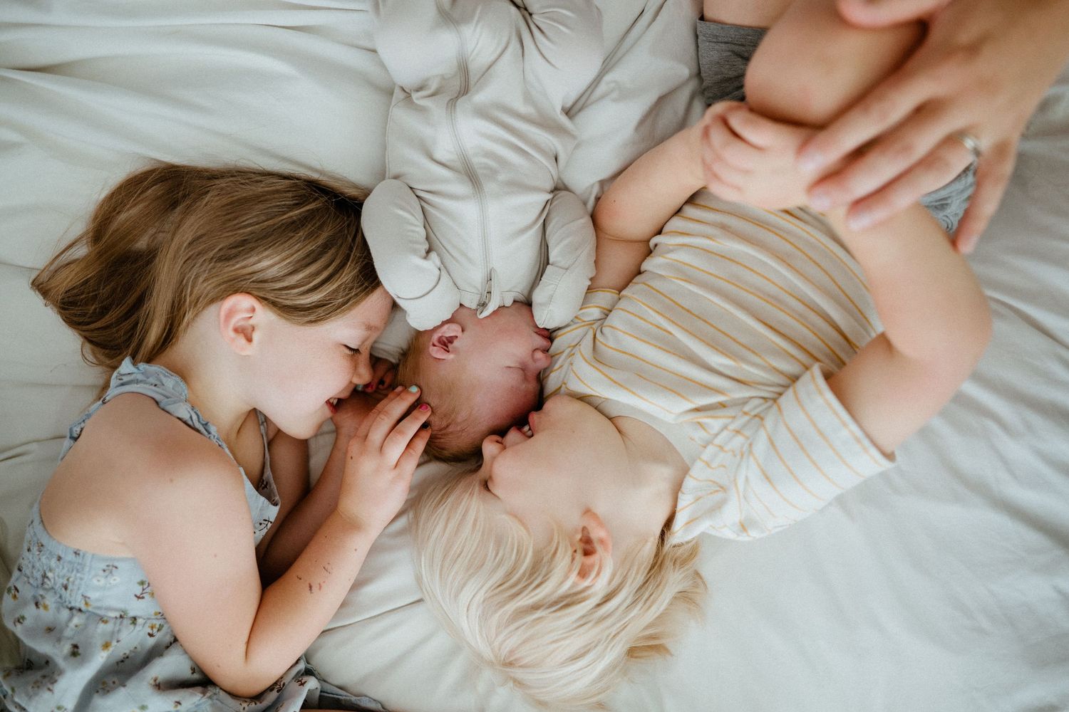Two young girls lying on a white bed lovingly gazing at their newborn sibling.
