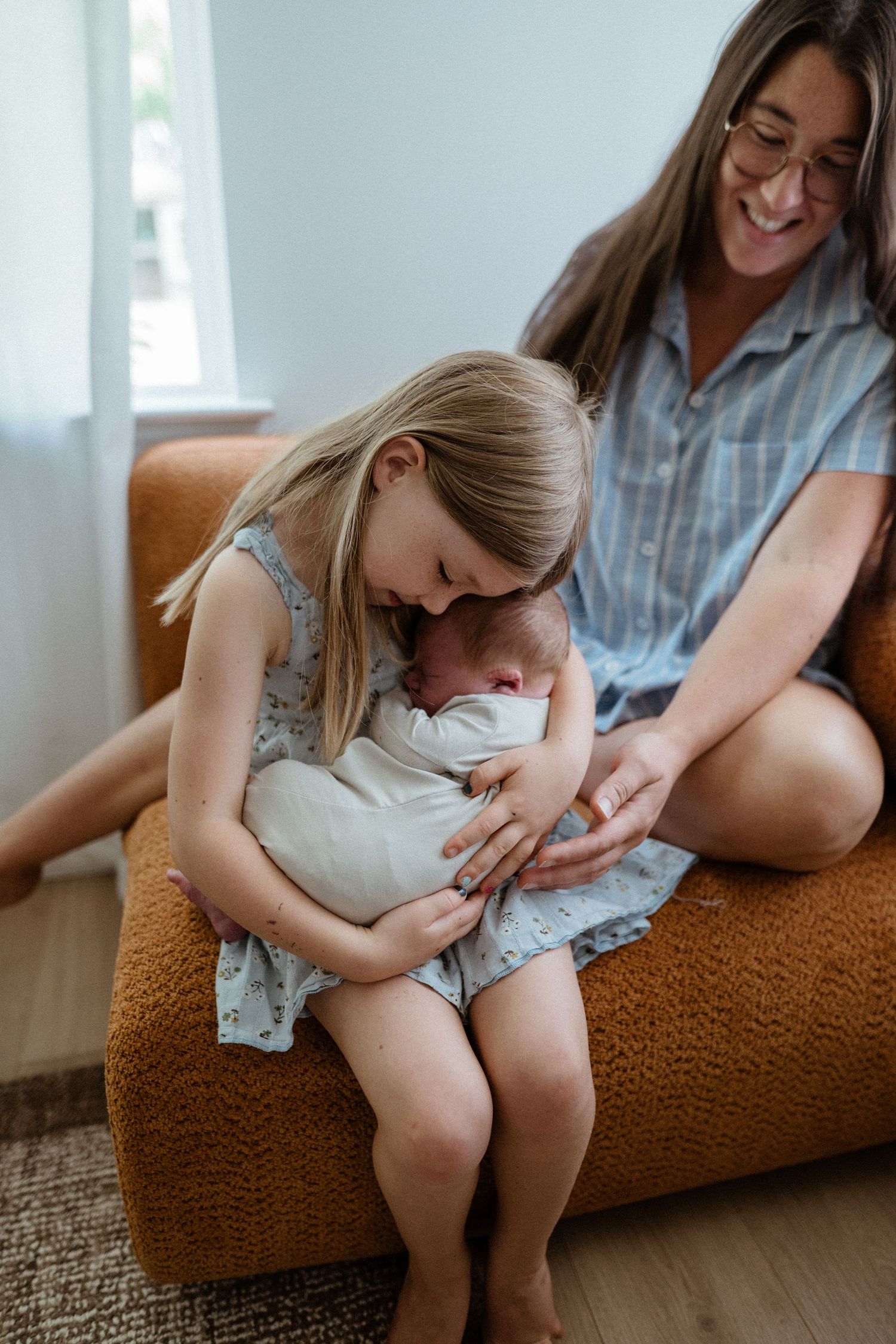 A smiling mother watches her young daughter lovingly hold a swaddled newborn on an orange chair.