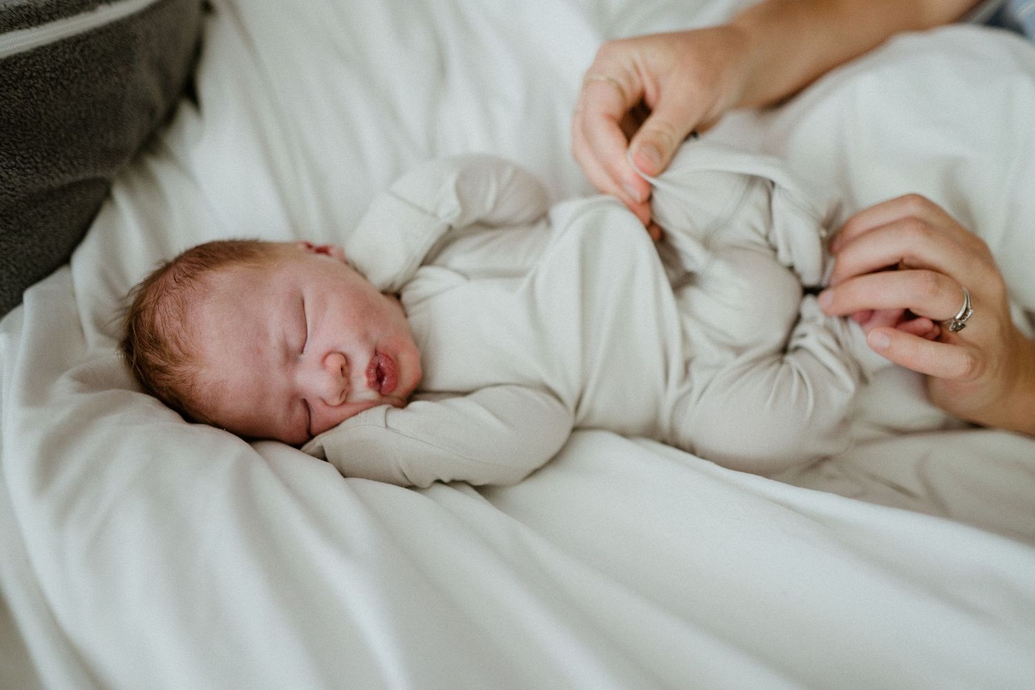 Newborn baby sleeping peacefully in white onesie on white bedding while parent gently touches tiny hand.