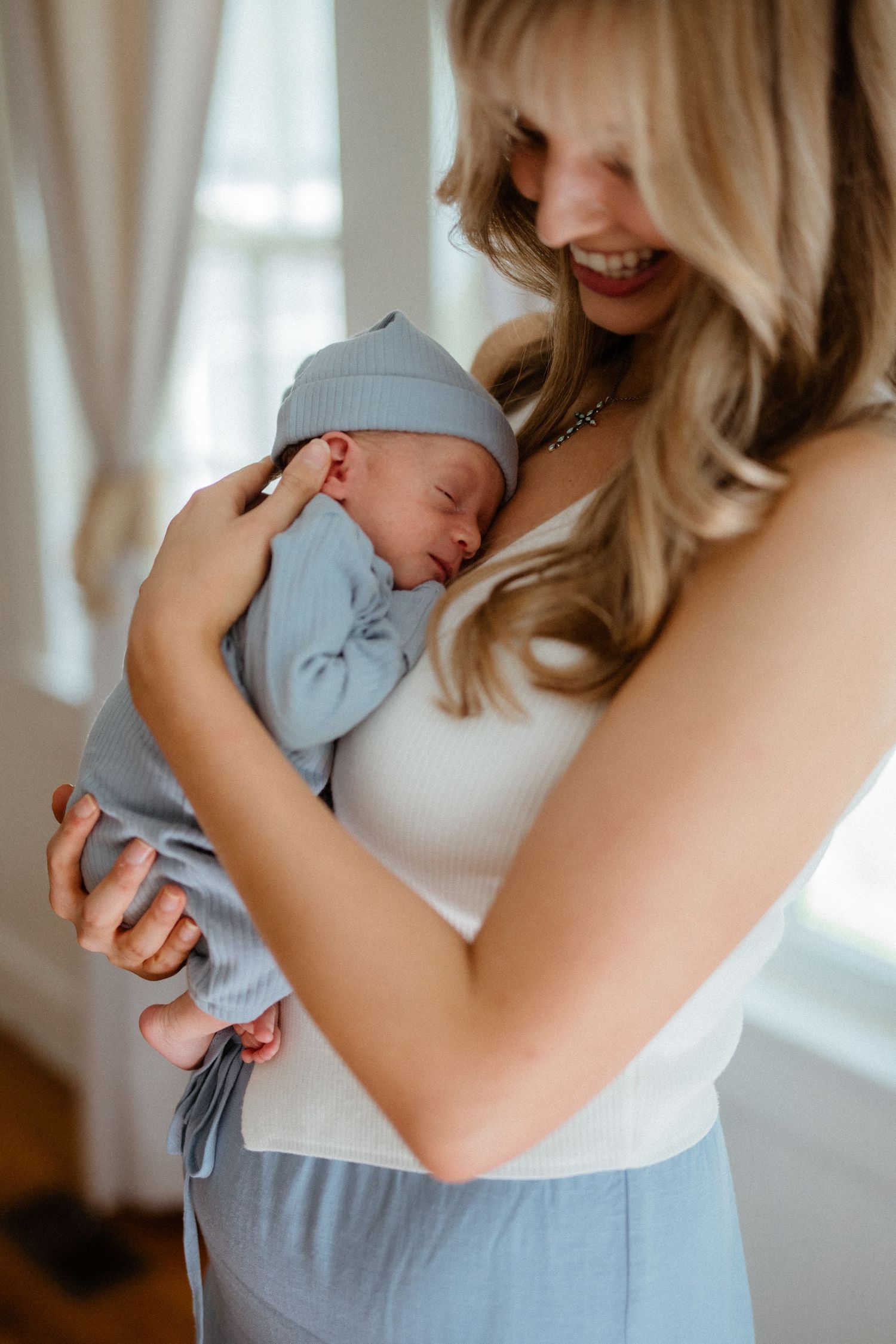 Smiling blonde mother gazing lovingly at sleeping newborn dressed in blue swaddle and knotted hat.