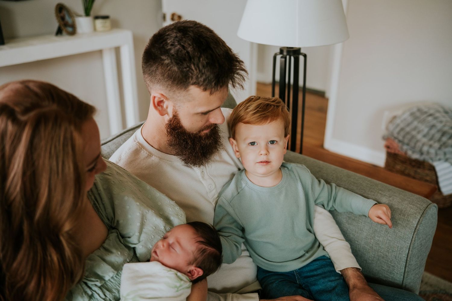 Bearded father and red-haired toddler boy look lovingly at sleeping newborn baby on a couch.