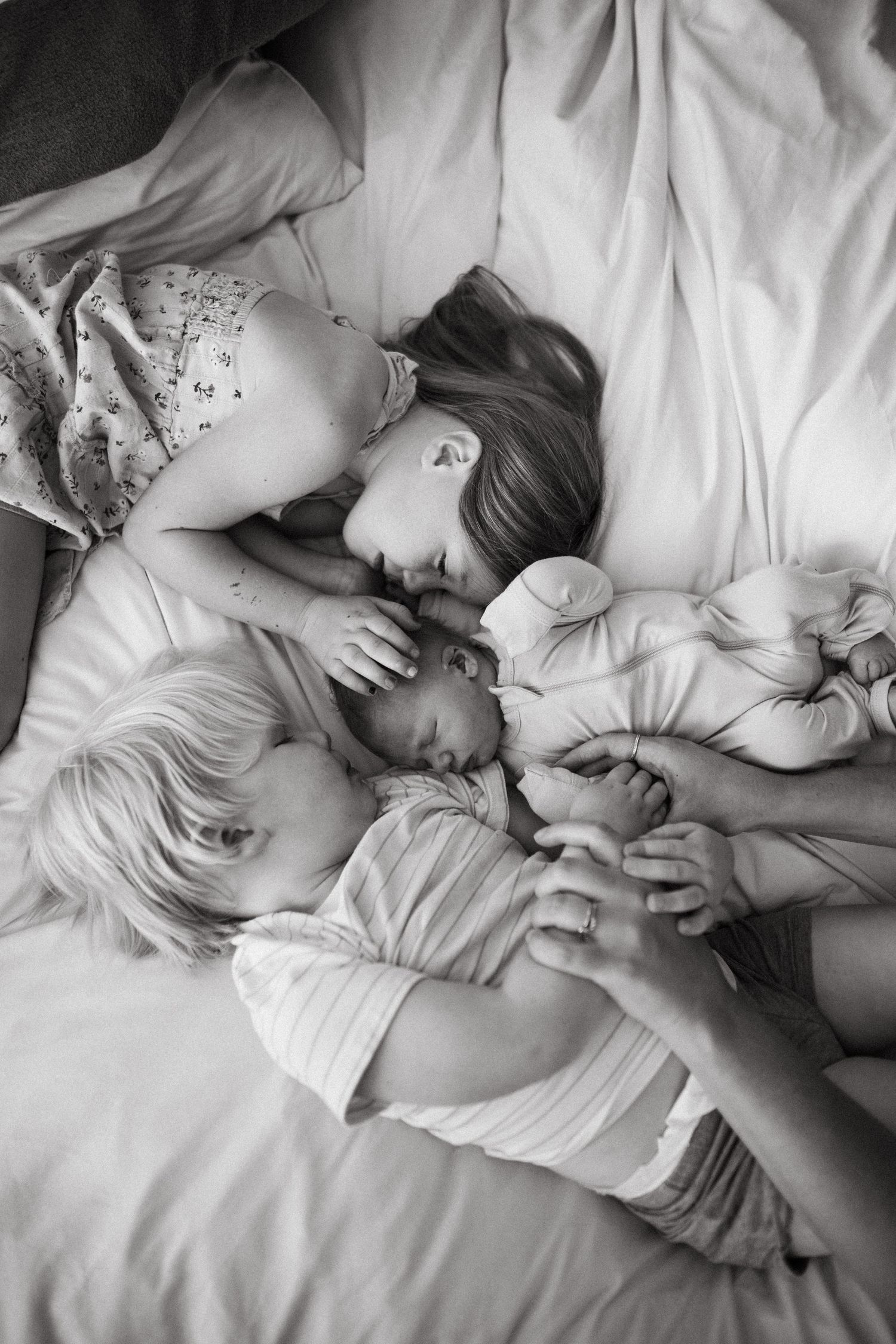 Black and white photo of two children tenderly embracing their sleeping newborn sibling on a bed.