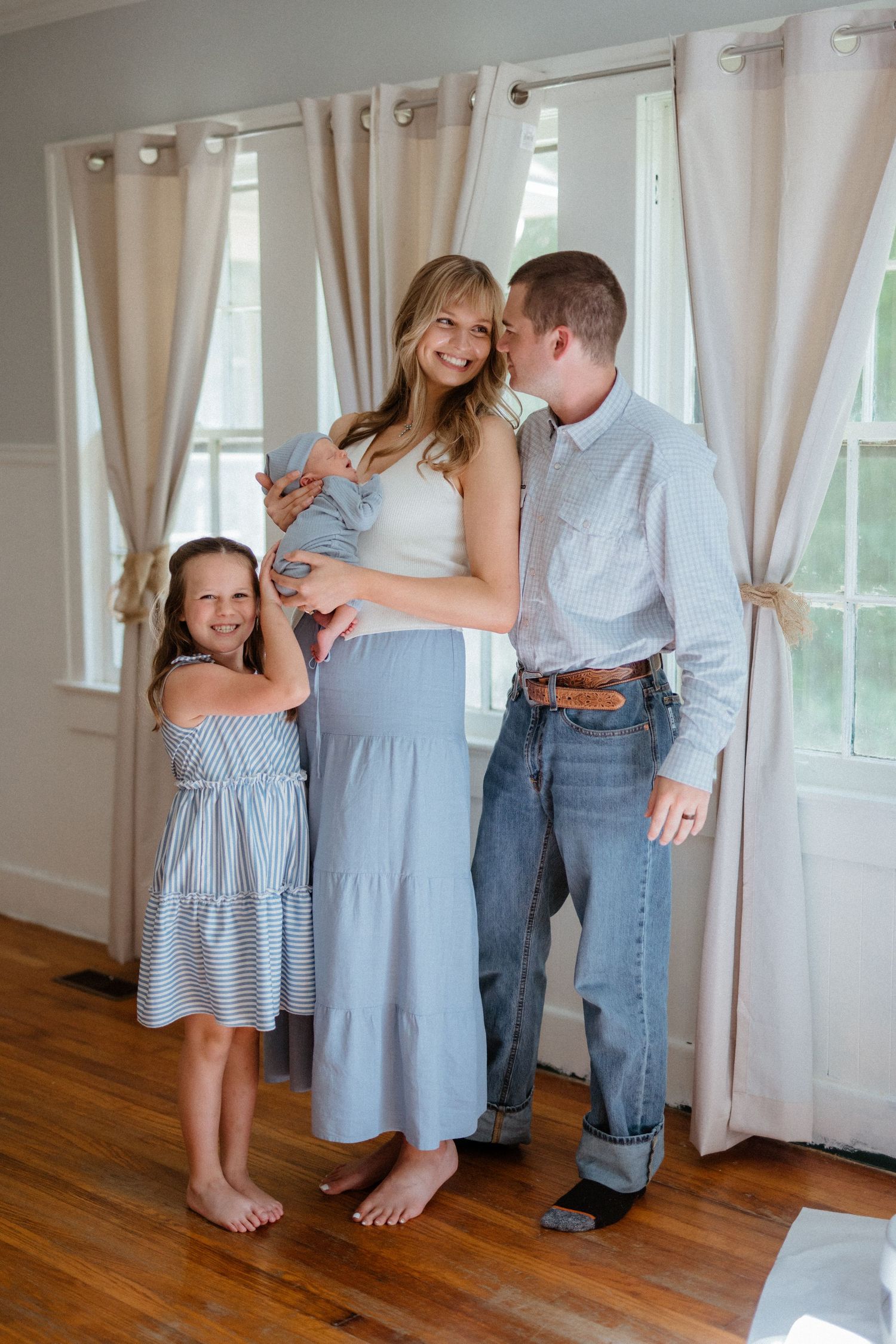Happy family portrait with mother holding newborn, father kissing her cheek, and young daughter smiling.