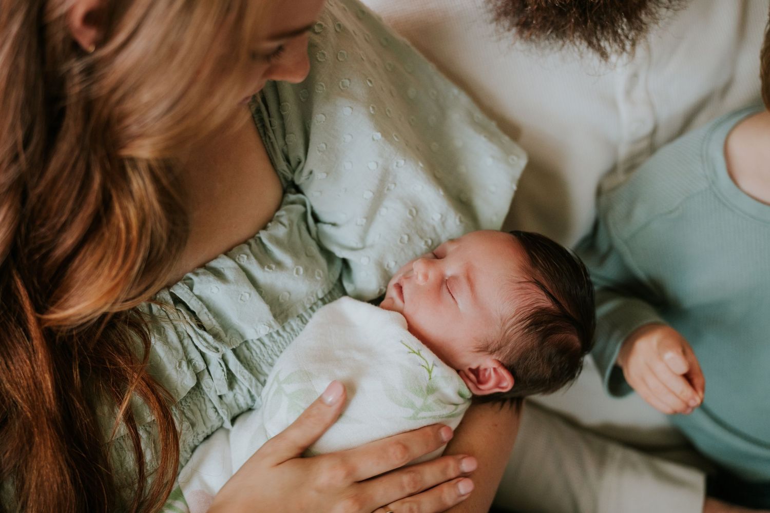 Close-up of mother in green dress cradling sleeping swaddled newborn baby with father nearby.
