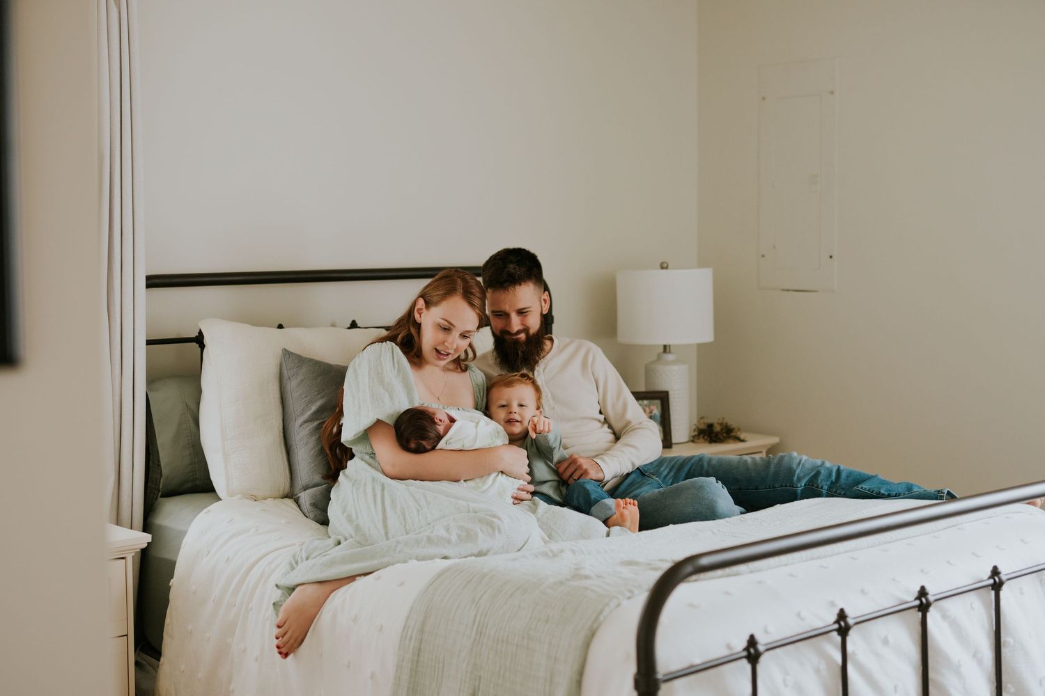 Loving family of four sitting on bed with newborn baby and toddler in bright bedroom.