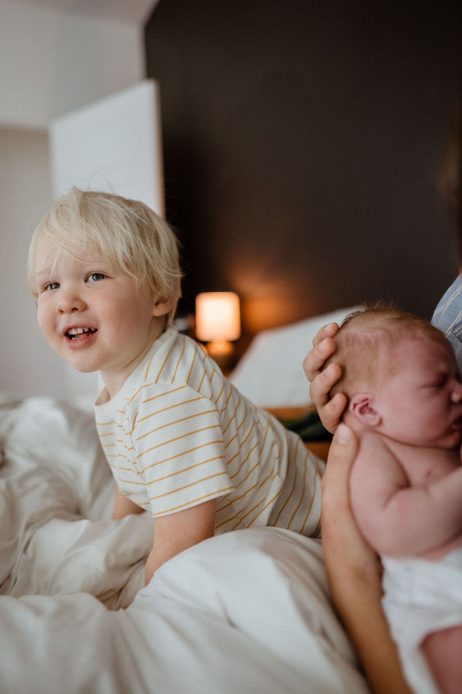 Happy blonde toddler in striped shirt sits on white bed next to newborn sibling.