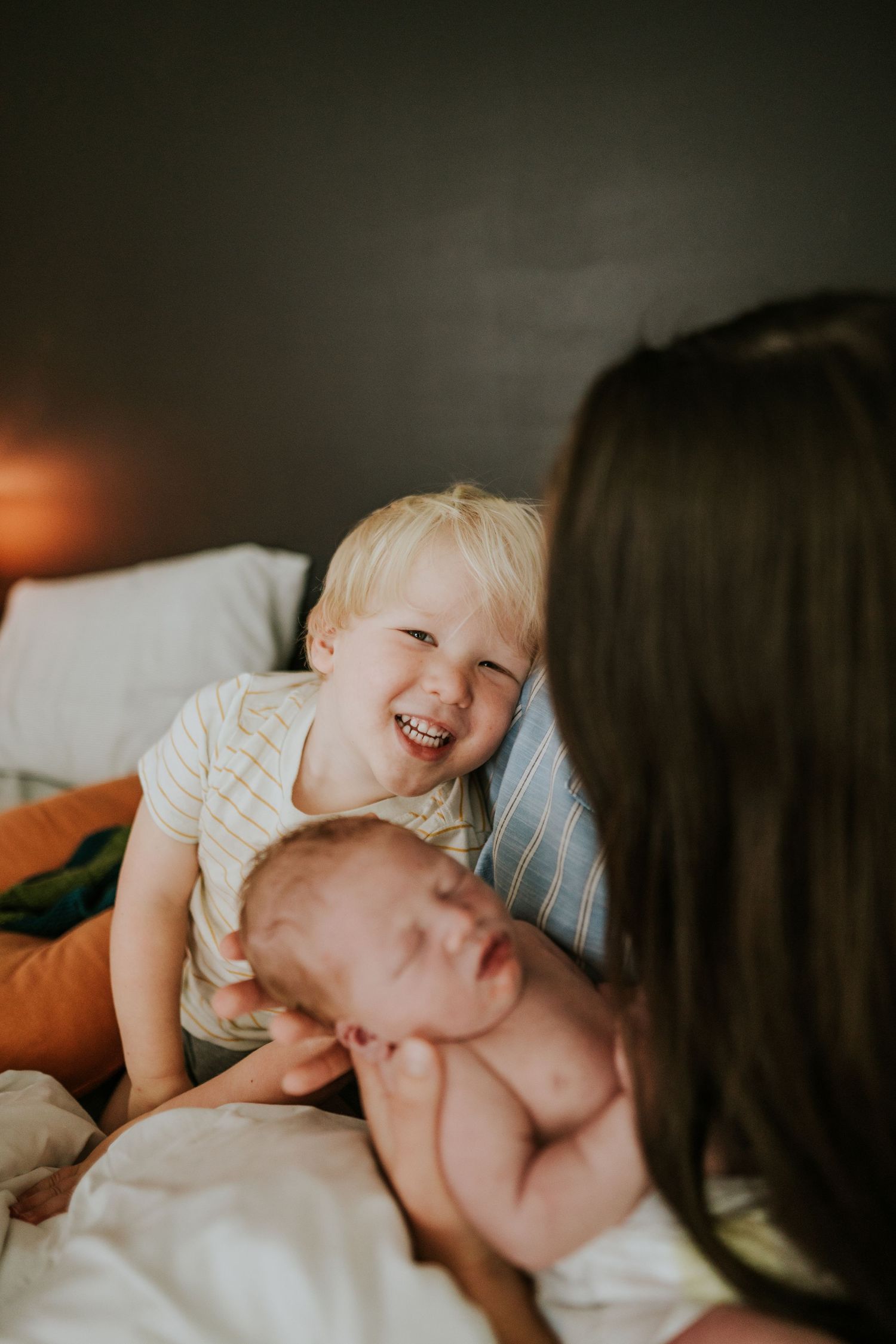 Laughing blonde toddler boy beside sleeping newborn baby held by mother on a bed.