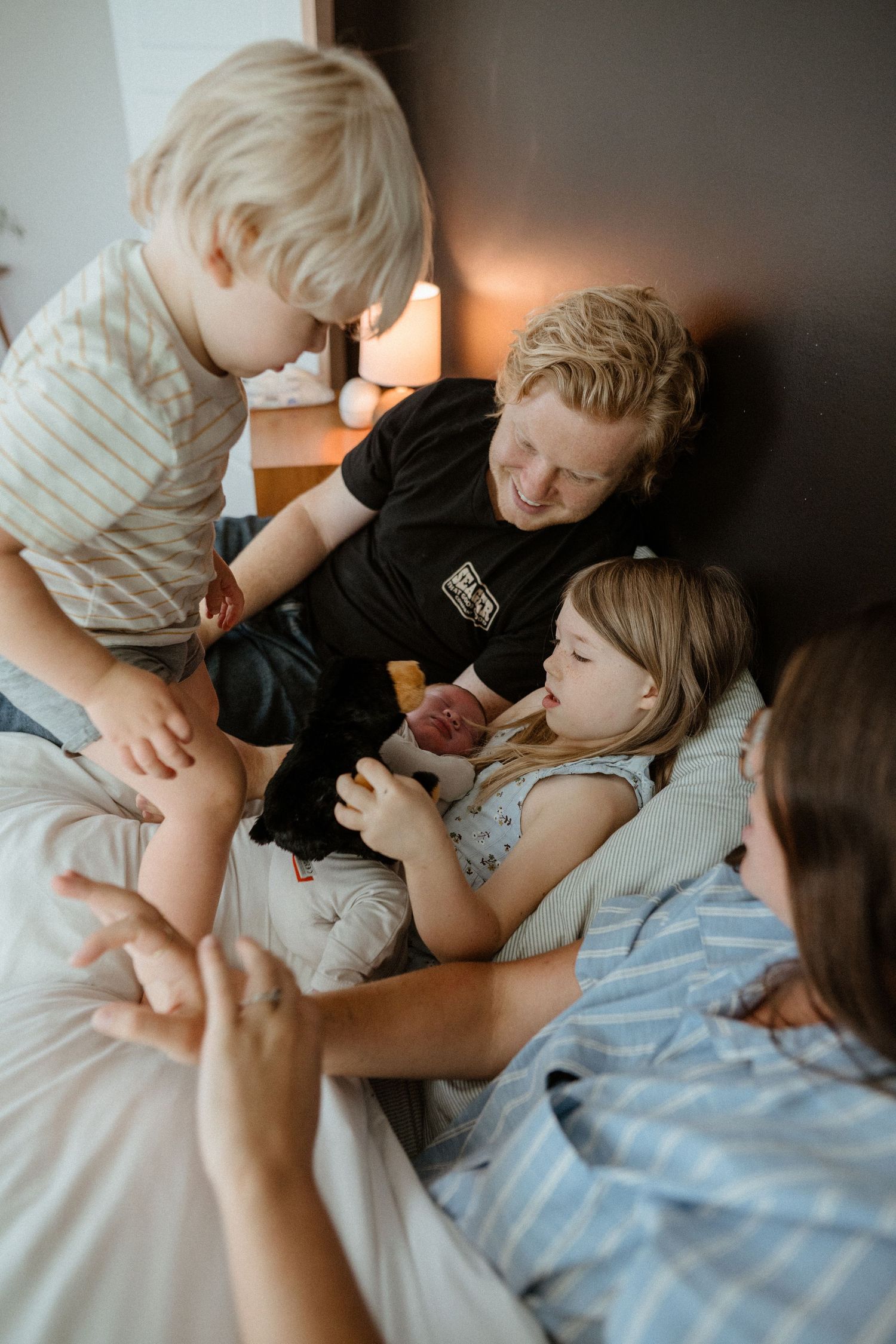 A father and two children gather on a bed, curiously looking at a newborn baby together.