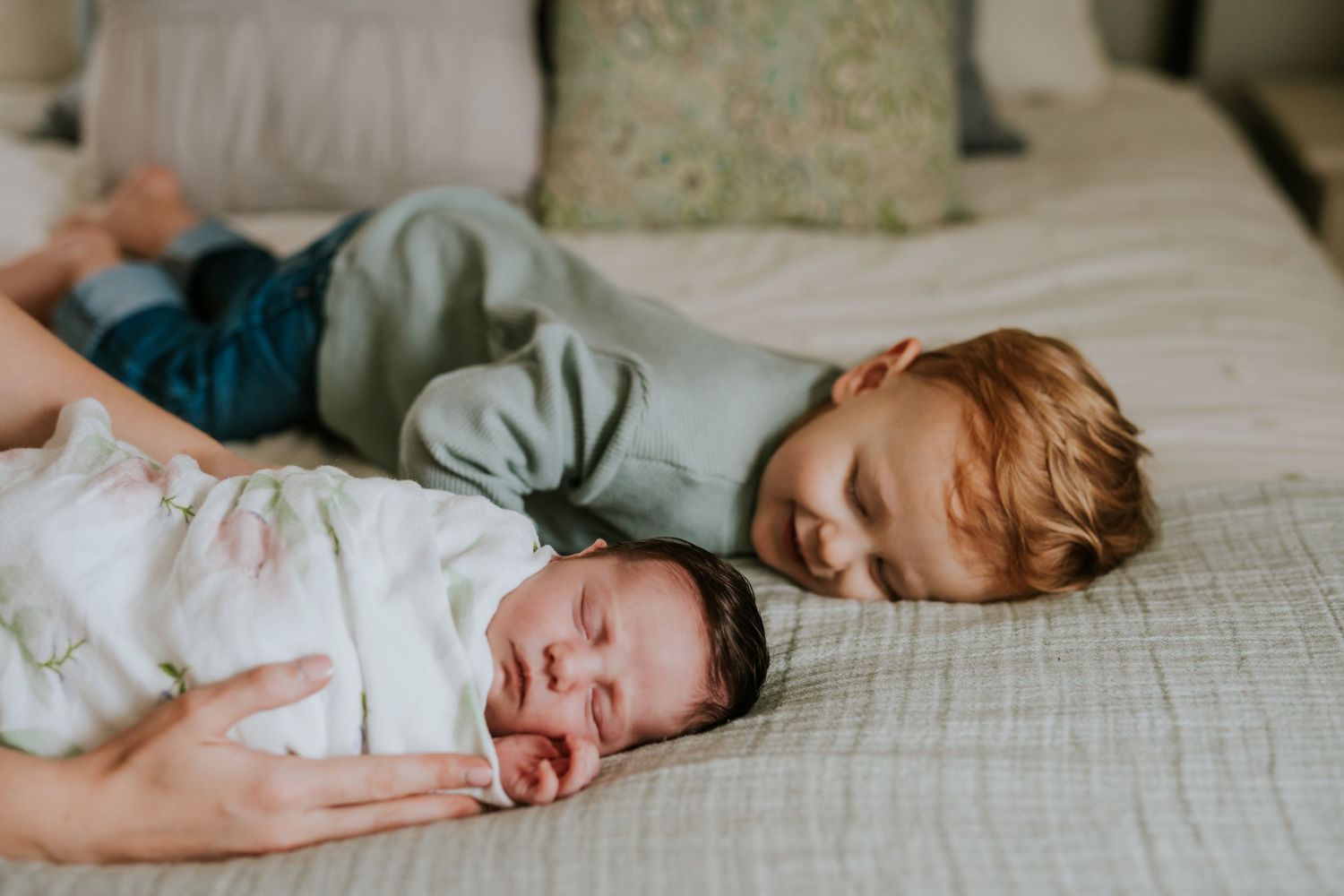 Toddler boy lying on bed beside sleeping swaddled newborn baby sibling.