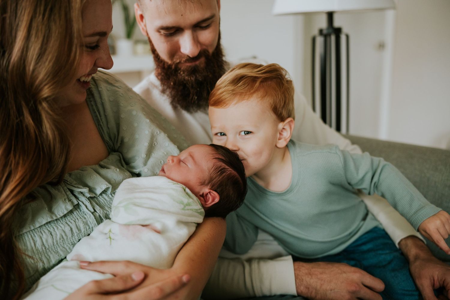 Red-haired toddler gently kisses sleeping swaddled newborn held by mother with father watching.